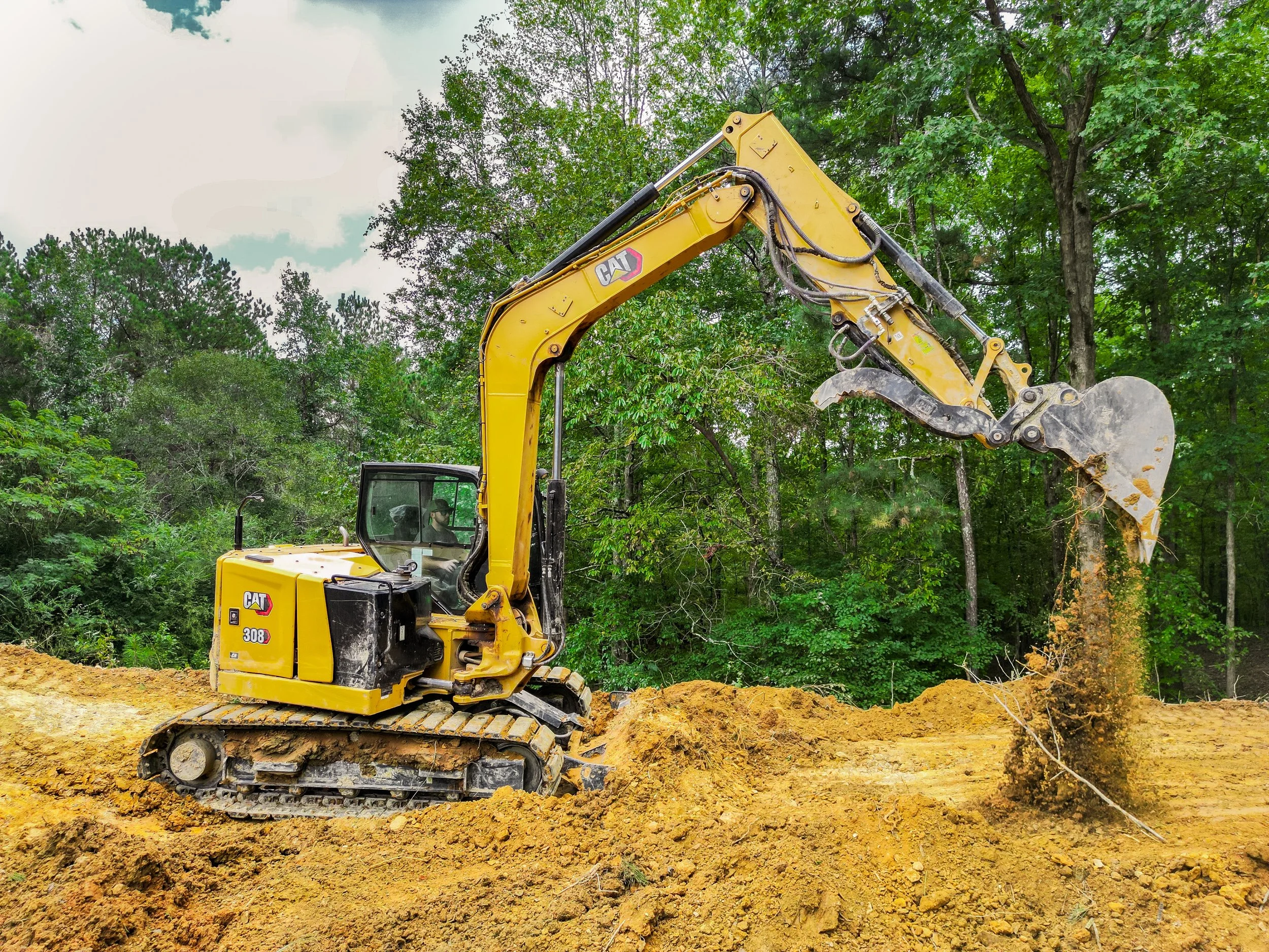 Yellow Caterpillar excavator digging into dirt at a construction site with green trees in the background.