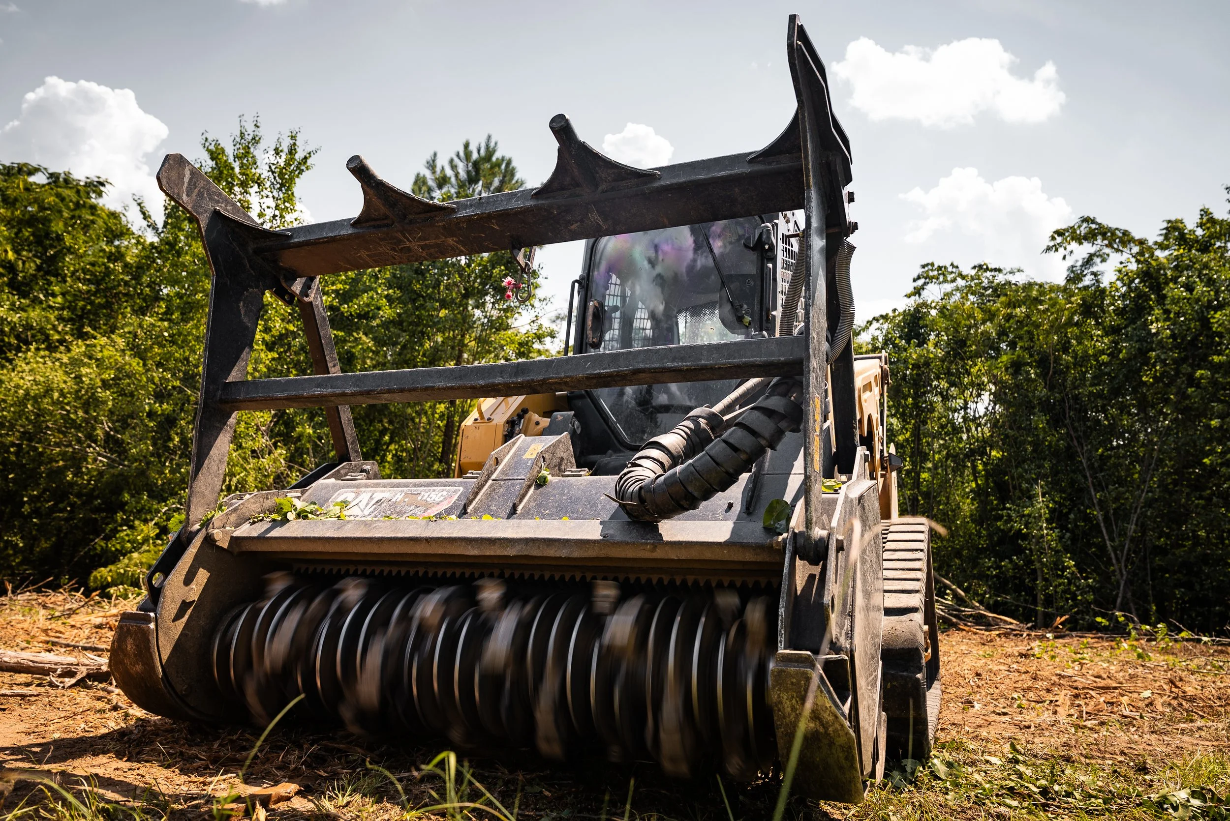 Front view of a heavy-duty skid steer loader with a rotating brush attachment working on cleared dirt with green trees in the background under partly cloudy sky.