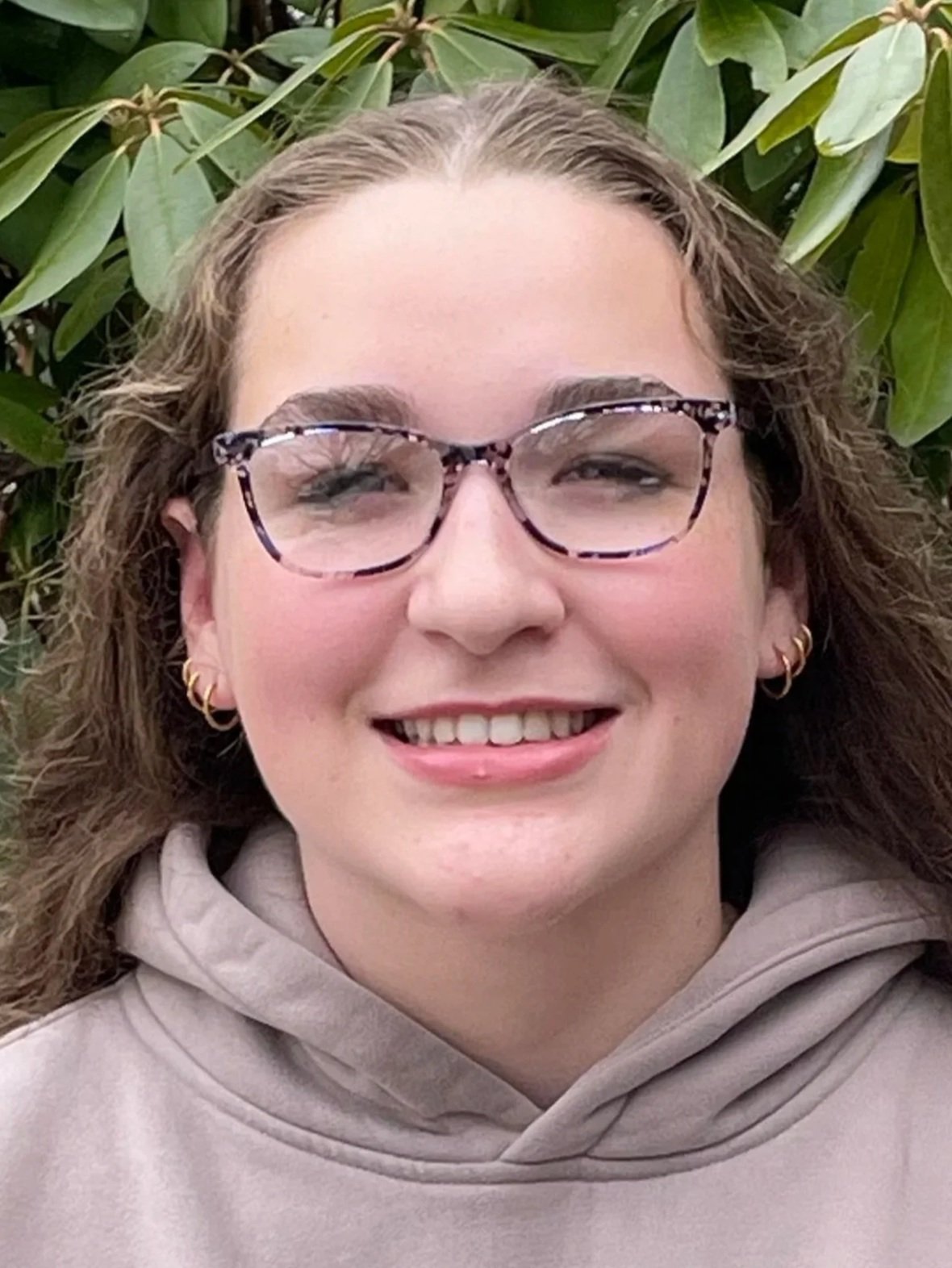 Close-up of a young woman smiling, wearing a light gray hoodie, outdoors with green foliage in the background.