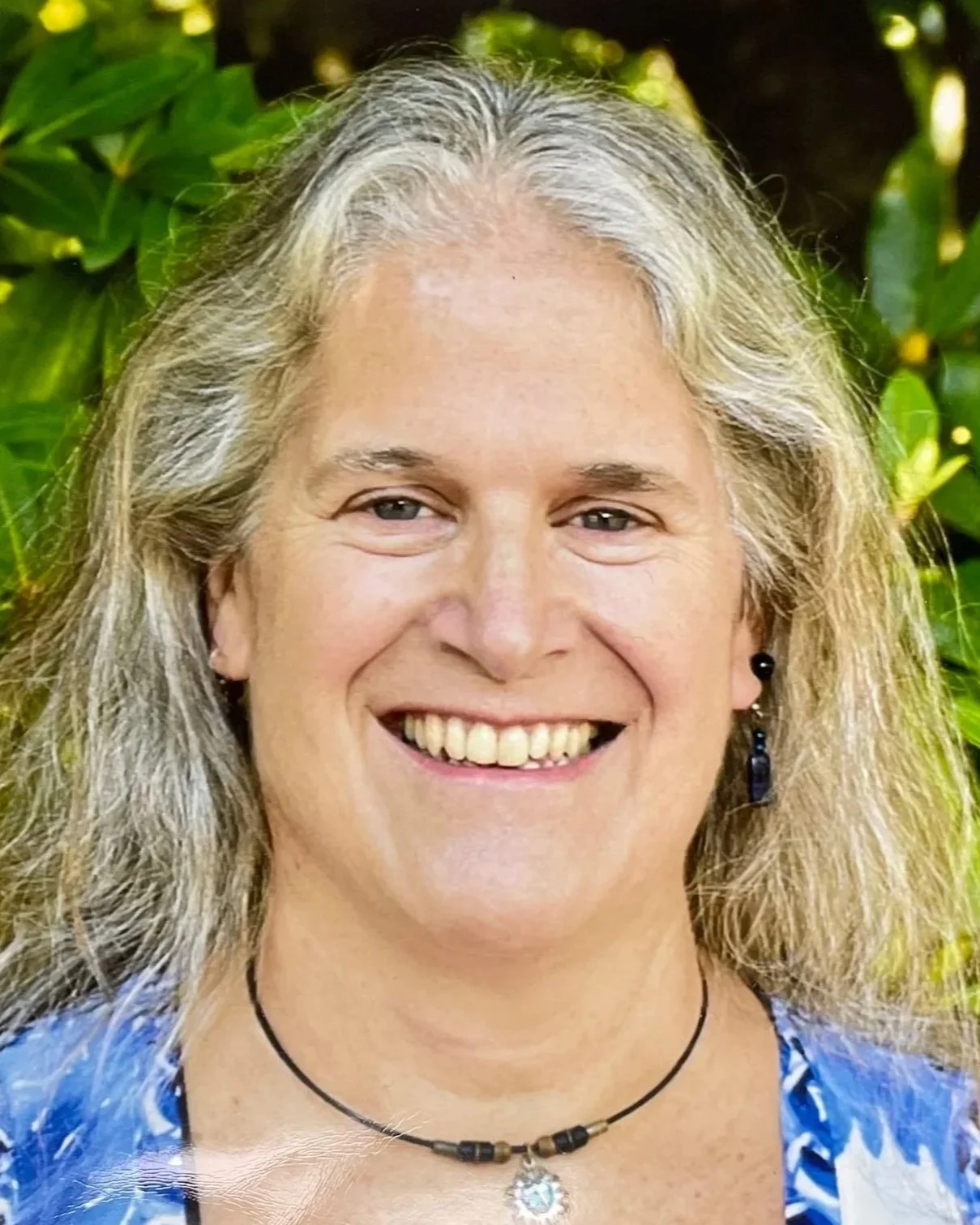 Close-up of a woman with curly gray hair and a warm smile, wearing a white turtleneck sweater, standing outdoors in front of green foliage.