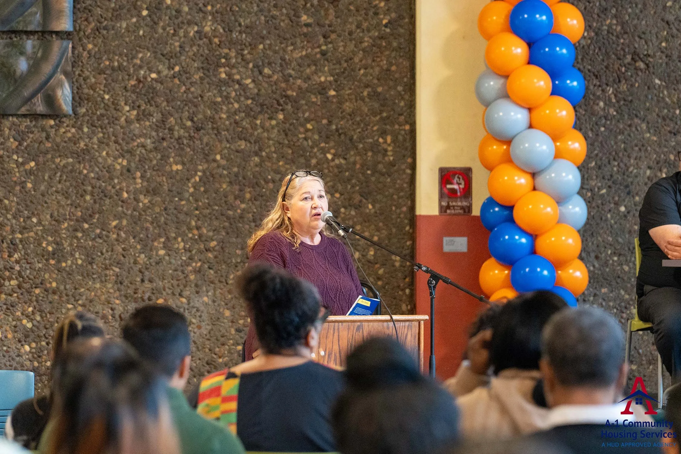 woman holding program for housing fair event and talking into microphone in front of a seated crowd