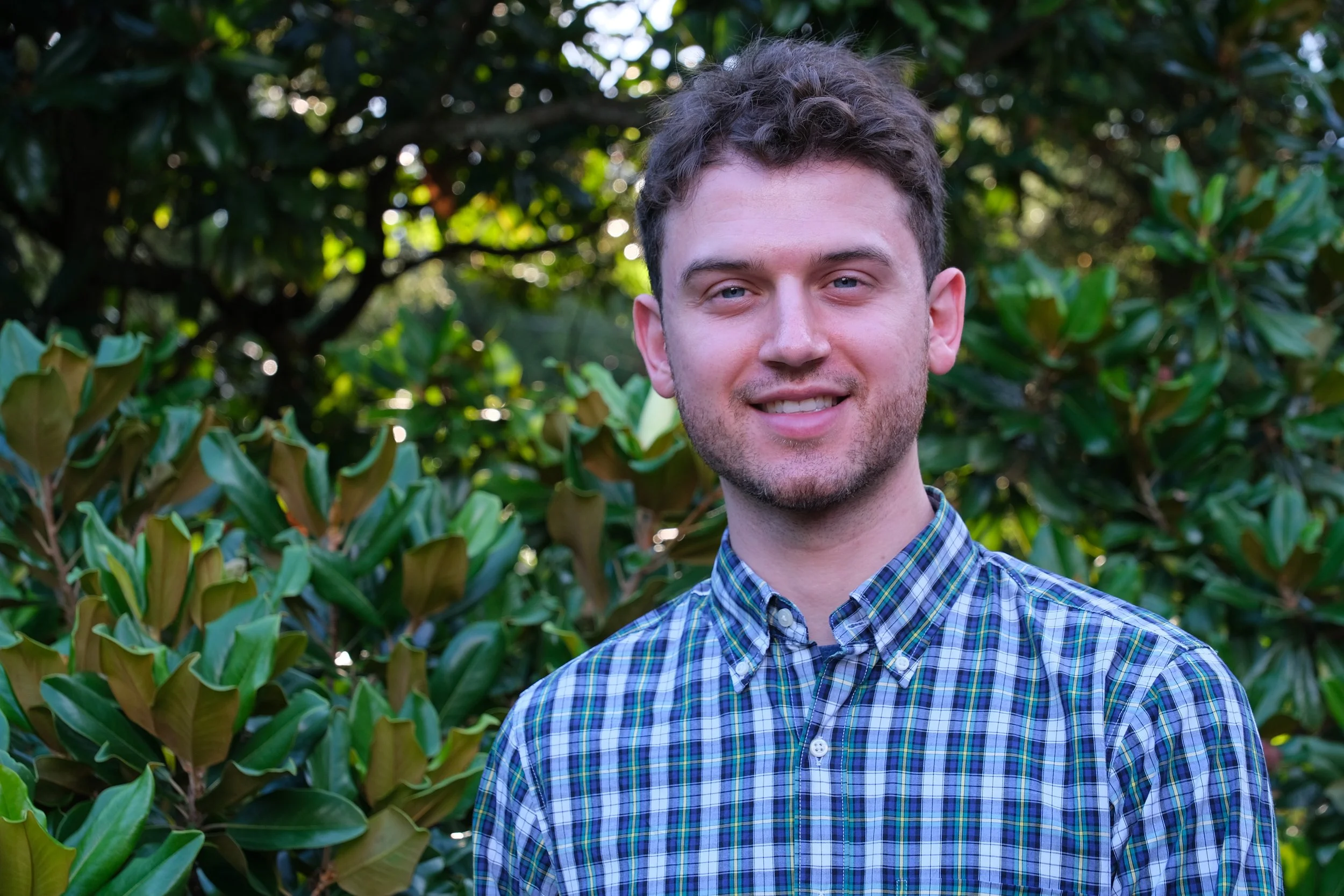 A young man with curly brown hair and blue eyes, wearing a blue plaid shirt, standing outdoors in front of green foliage and trees.