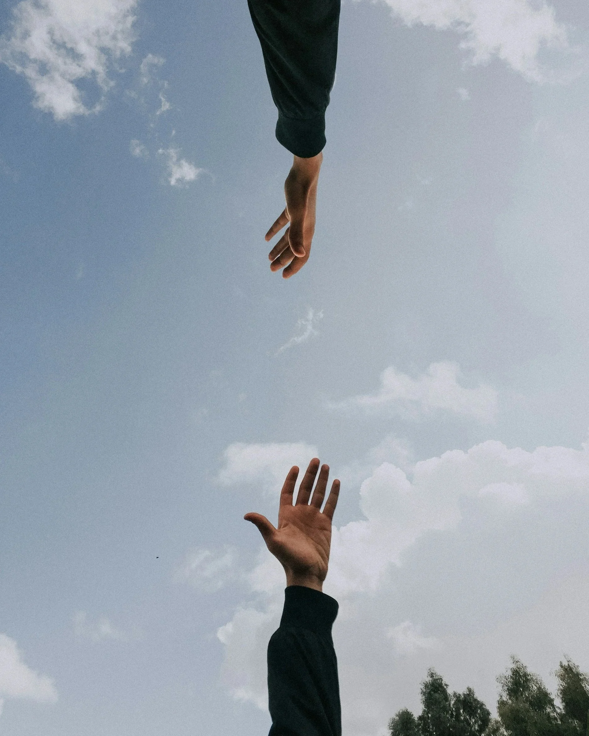 Two hands reaching towards each other in the sky, one higher than the other, against a backdrop of clouds and trees.