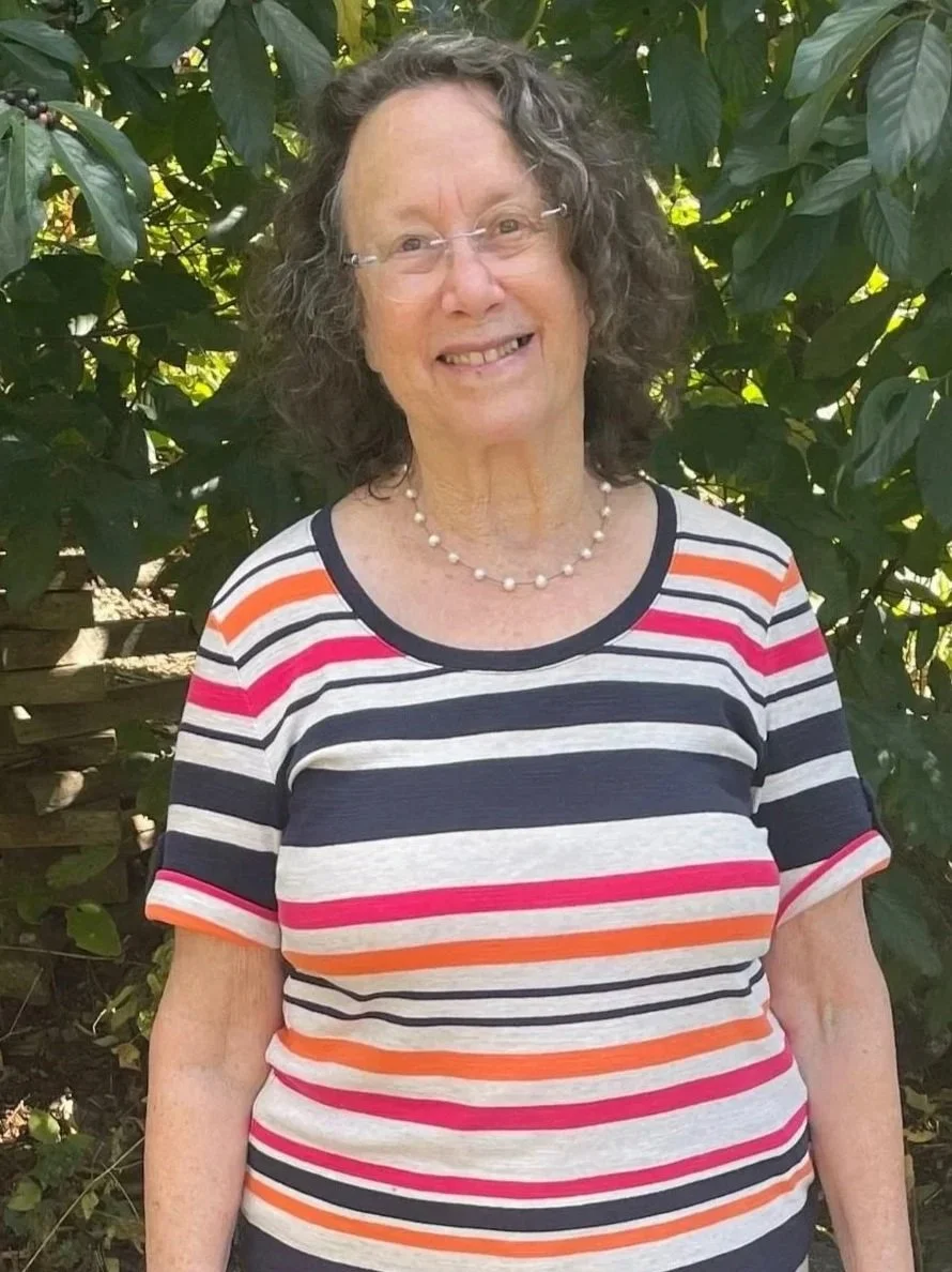 An elderly woman with curly gray hair, glasses, and a pearl necklace, smiling outdoors in front of green foliage, wearing a colorful striped shirt.