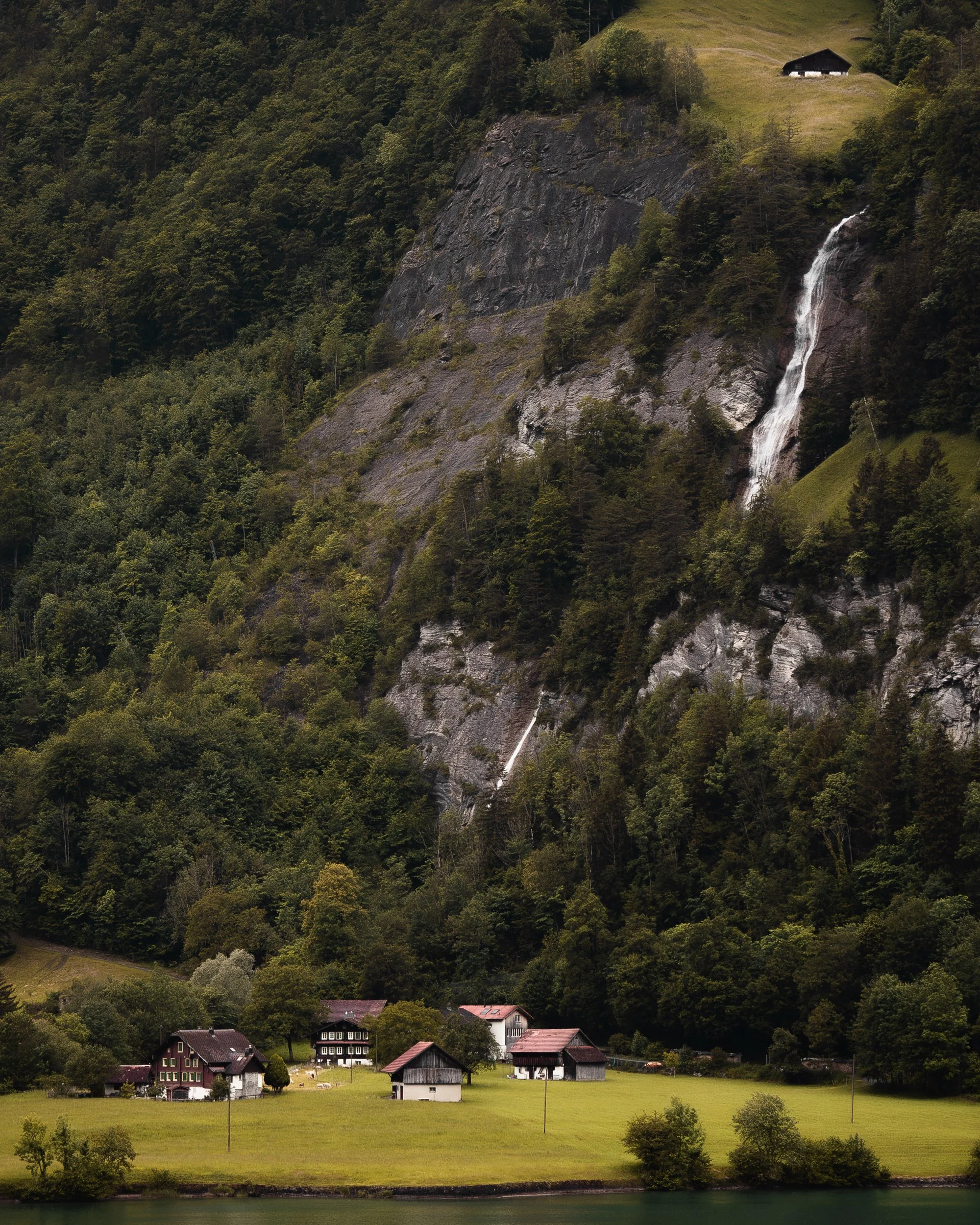 Alpine waterfall cascading down a steep mountainside above a traditional Swiss village surrounded by forest.