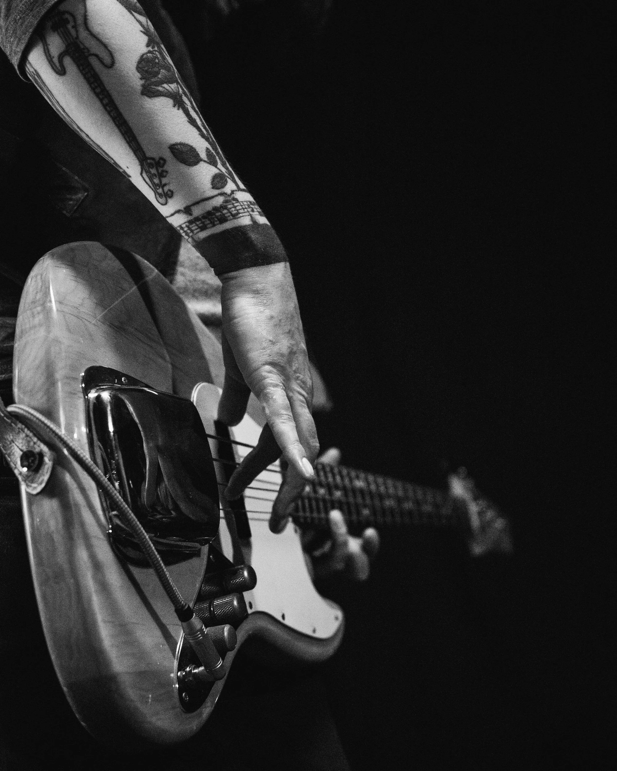 Black and white close-up of tattooed arm playing bass guitar during rehearsal, atmospheric music portrait, photography by Jerre Maier.