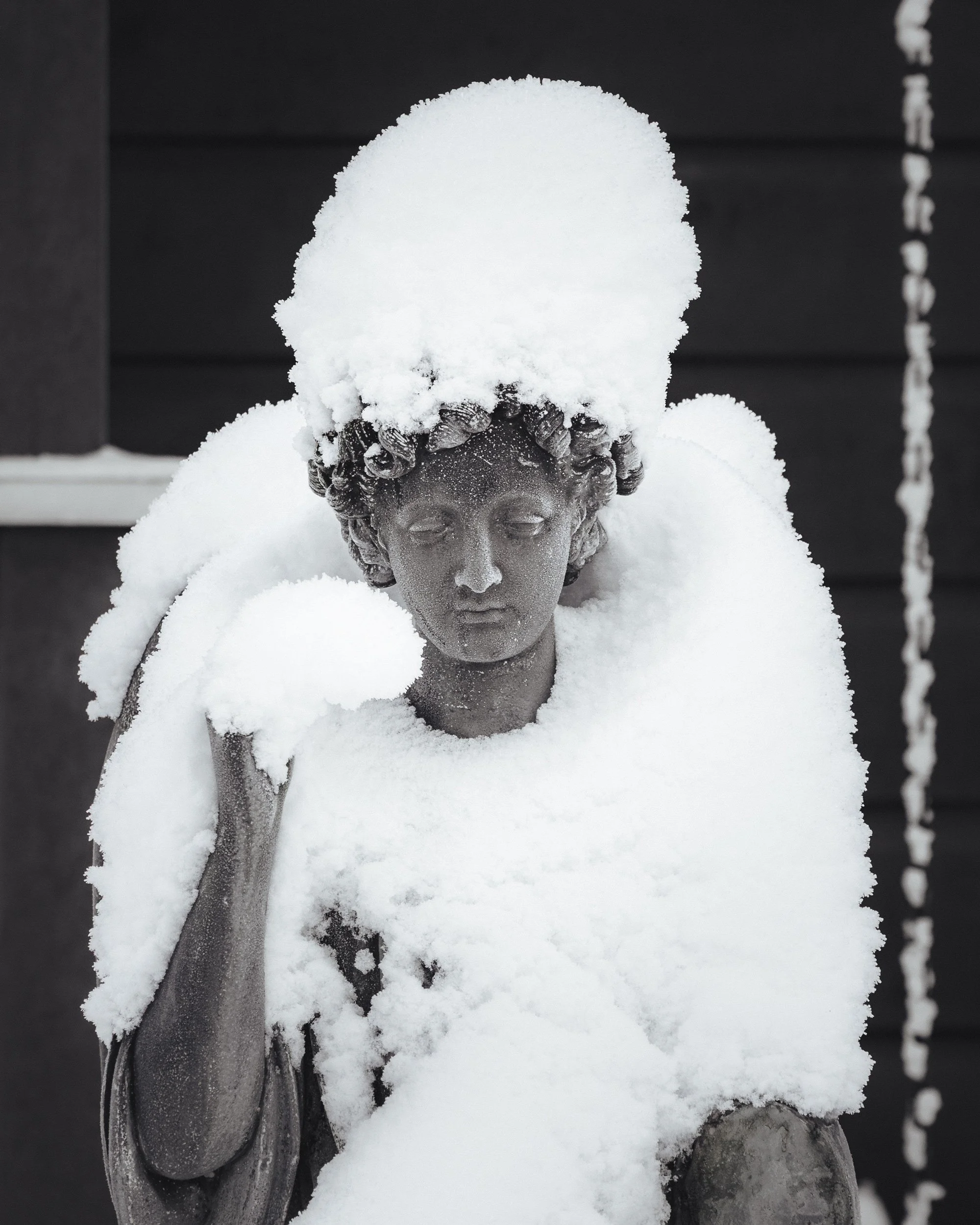 Stone angel statue covered in fresh snow, serene winter scene with soft monochrome tones.