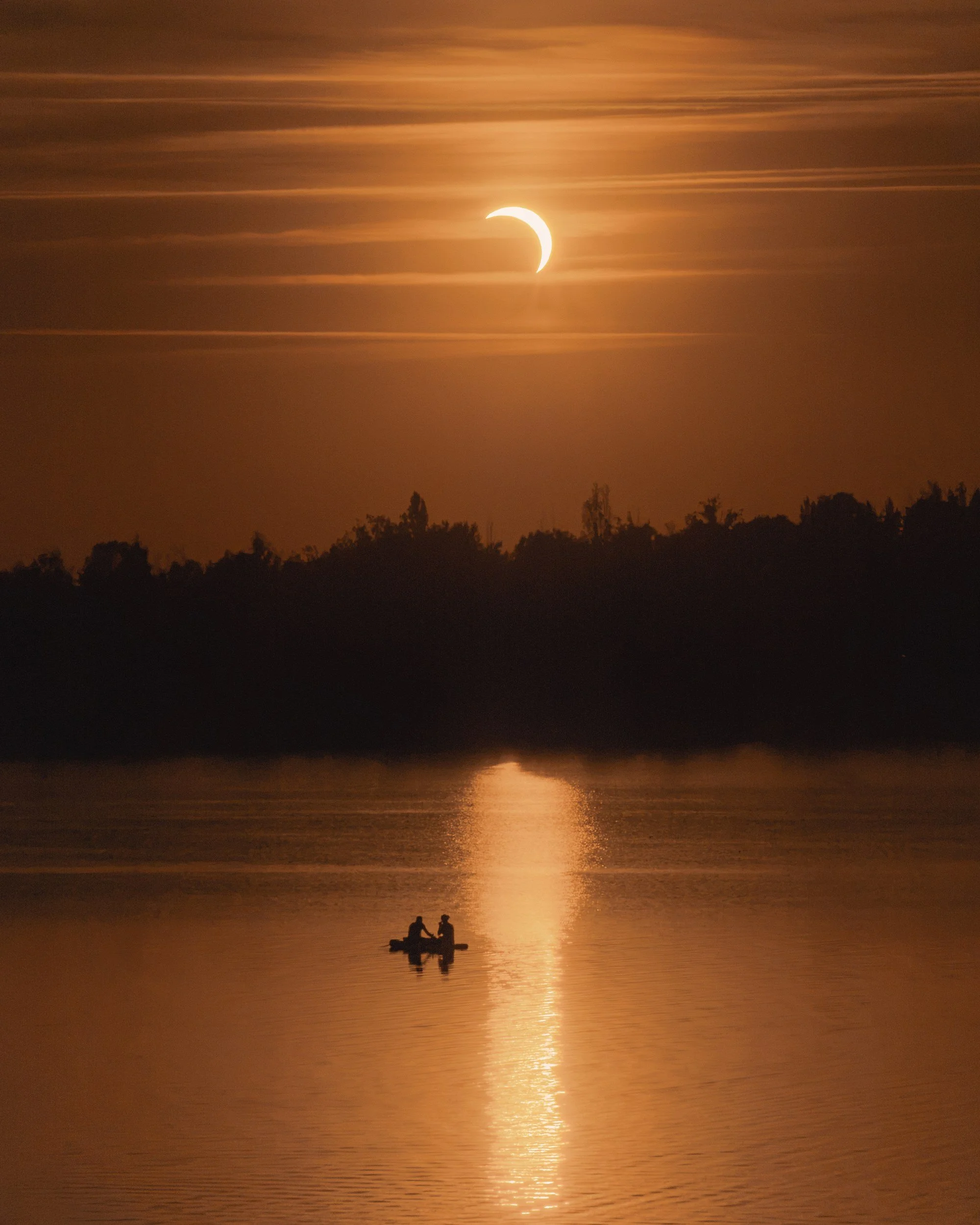A partial solar eclipse glows above a lake while two paddlers cross the sunlit reflection.
