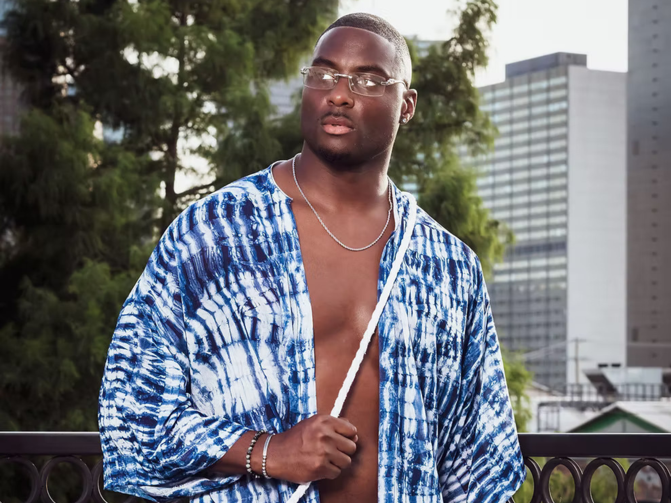 A young man wearing glasses and a blue patterned unbuttoned shirt, standing outdoors near a black fence with green trees and tall buildings in the background.