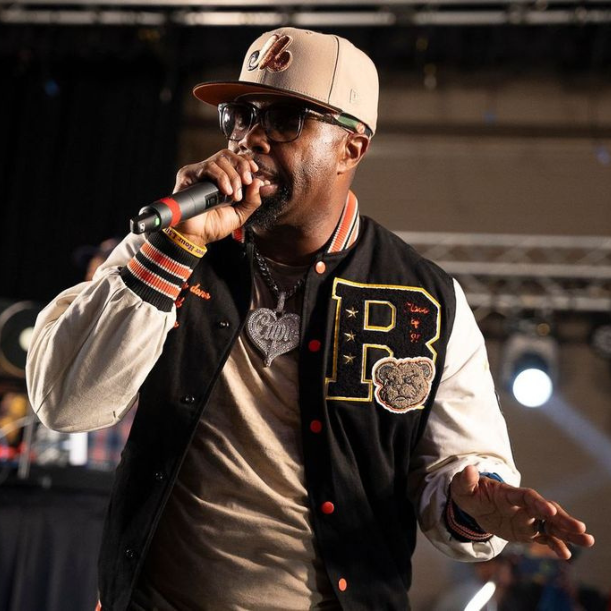Close-up of a man wearing a brown New York Yankees cap, black glasses, a fur-lined vest, a denim jacket, and a black t-shirt with white text, holding a microphone up to his mouth, with blurred people in the background.