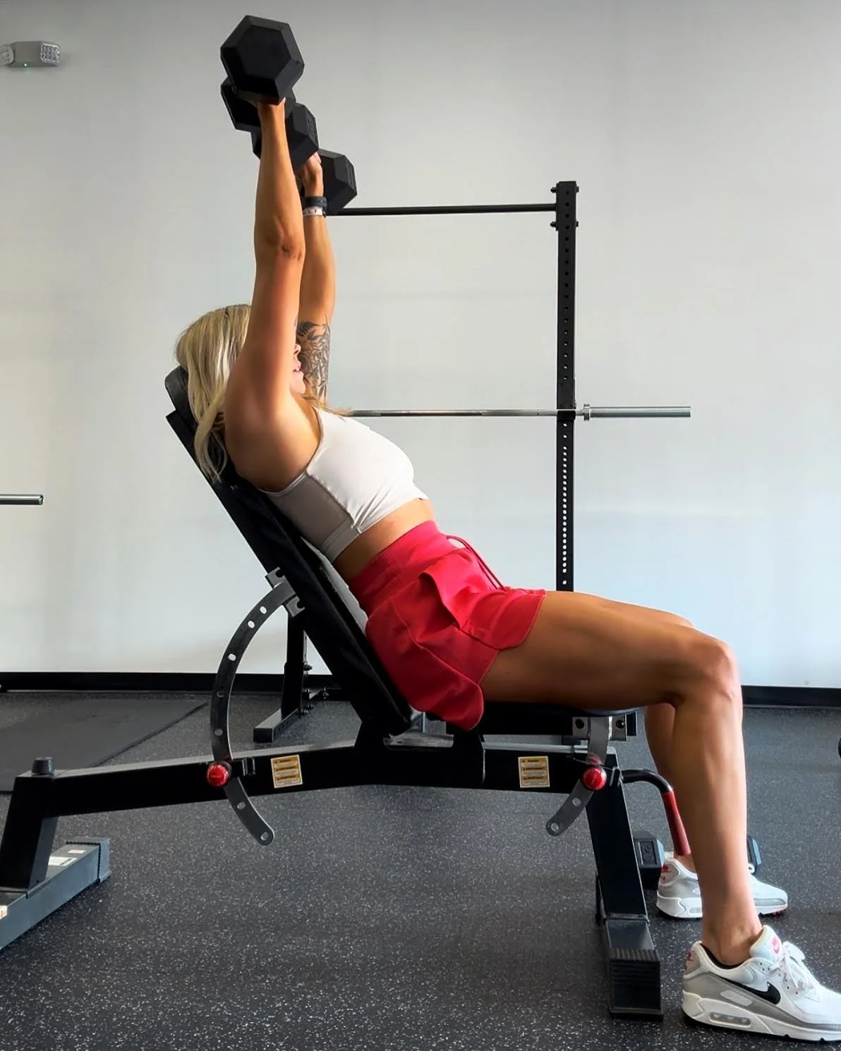 A woman with blonde hair and tattoos on her arm performing a seated dumbbell shoulder press exercise on a gym bench, with a barbell rack in the background.