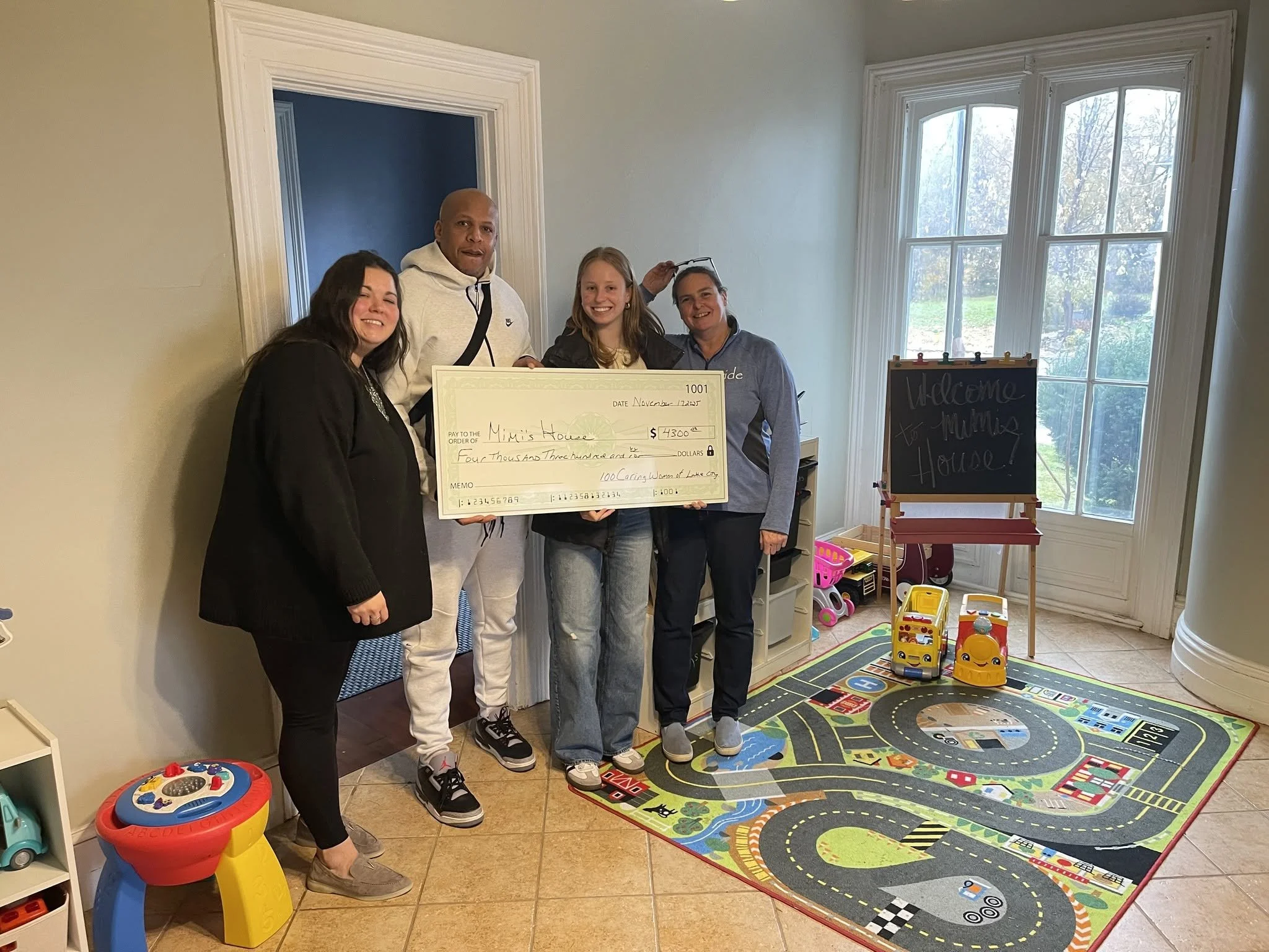 Group of four people standing inside a room with a colorful children's play area, including a small play kitchen, a activity table, and a play rug with roads and buildings. They are holding a large check awarded to Mimis House.
