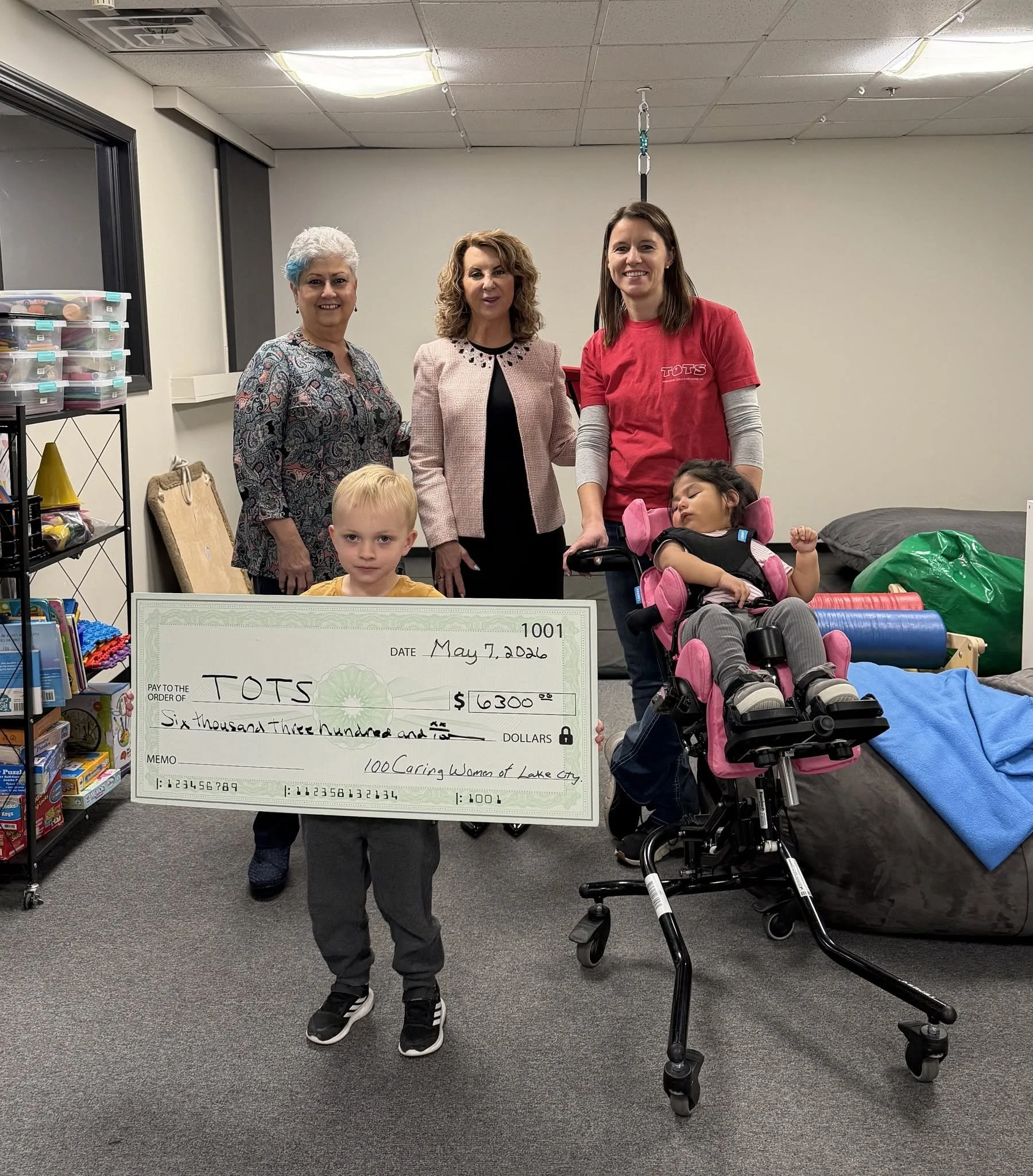 Four women and two children posing indoors, one of the children sitting in a stroller, holding a large check for $6,300 made out to TOTS, with a date of May 7, 2026. Behind them are store shelves, a window, and some equipment.