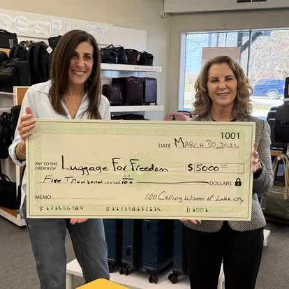 Two women smiling and holding a large oversized check for $5,000 made out to 'Luggage for Freedom' in an office setting.