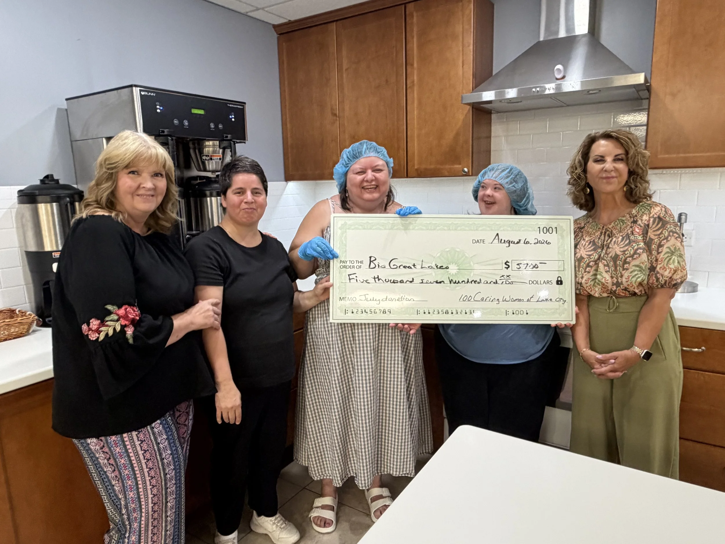 Group of five women standing in a kitchen, holding a large check made out to Big Great Lakes for $5300, dated August 6, 2020, representing a July donation from 100 Caring Women of Lake County. The women are smiling and wearing casual clothing and hair coverings.