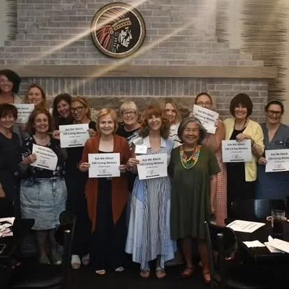 Group of women standing together in a room, holding certificates and smiling, with a U.S. Army insignia above them on the wall.