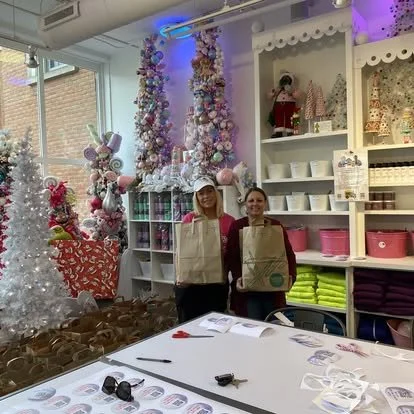 Two women standing in a store decorated with Christmas trees, ornaments, and holiday decorations, smiling while holding shopping bags, with a table and holiday items around them.