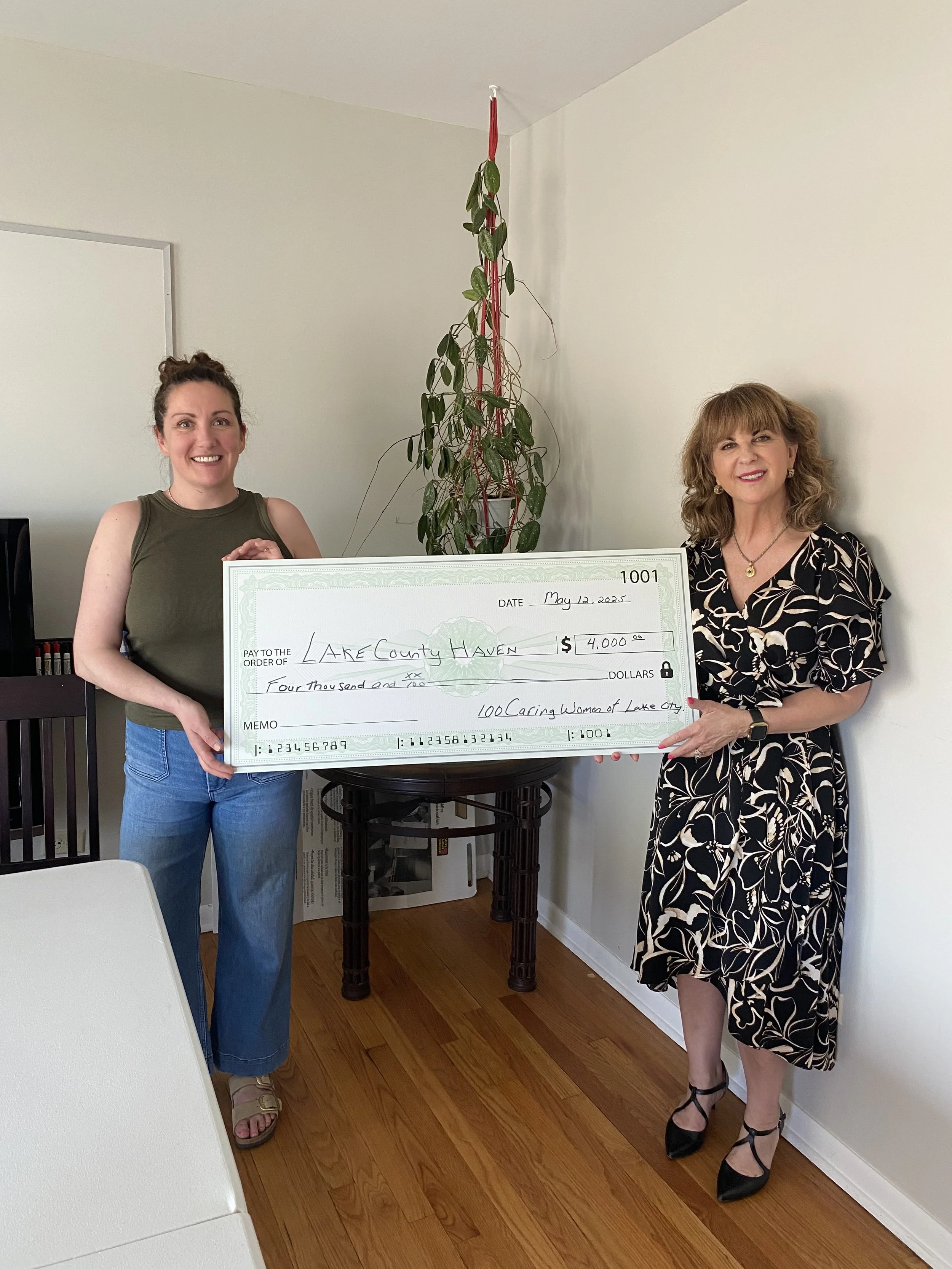 Two women standing in a room, holding a large ceremonial check for $4,000 made out to Lake County Haven, dated May 12, 2023. The woman on the left wears a green sleeveless top and blue jeans, smiling. The woman on the right wears a patterned black and white dress and black heels, also smiling. There is a tall potted plant between them and a white wall in the background.