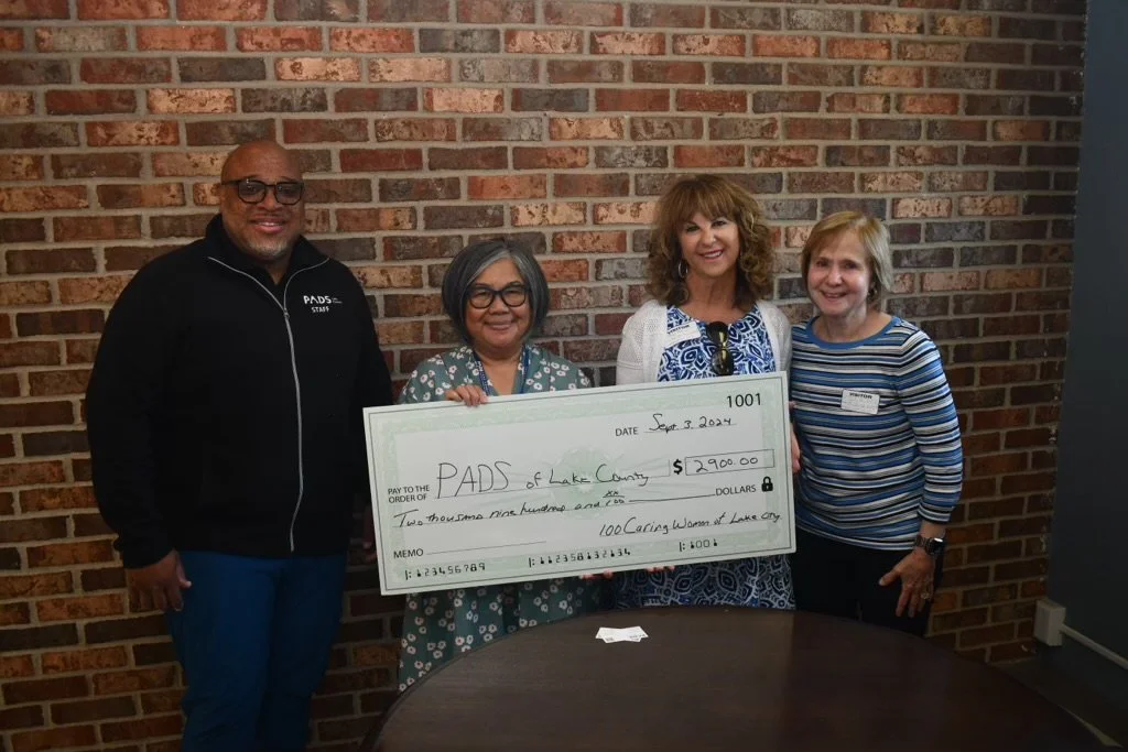 Four people standing together, holding a large check for $2,900 made out to PADS of Lake County, with a brick wall in the background.