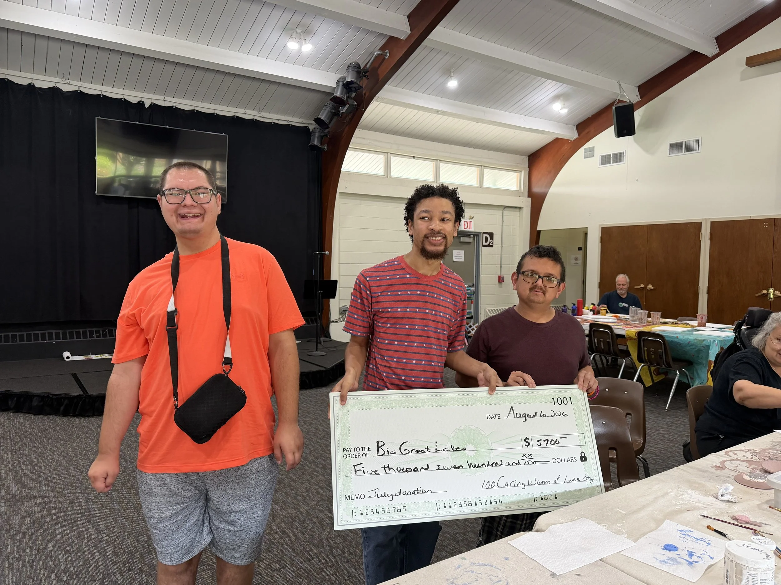 Three men standing in a room, one holding a large check for $5700. The man on the left is wearing glasses and an orange shirt, the man in the middle is wearing a striped shirt, and the man on the right is wearing a maroon shirt. There are tables with items and a woman seated at the table on the right. The background shows a stage, a TV, and large windows.