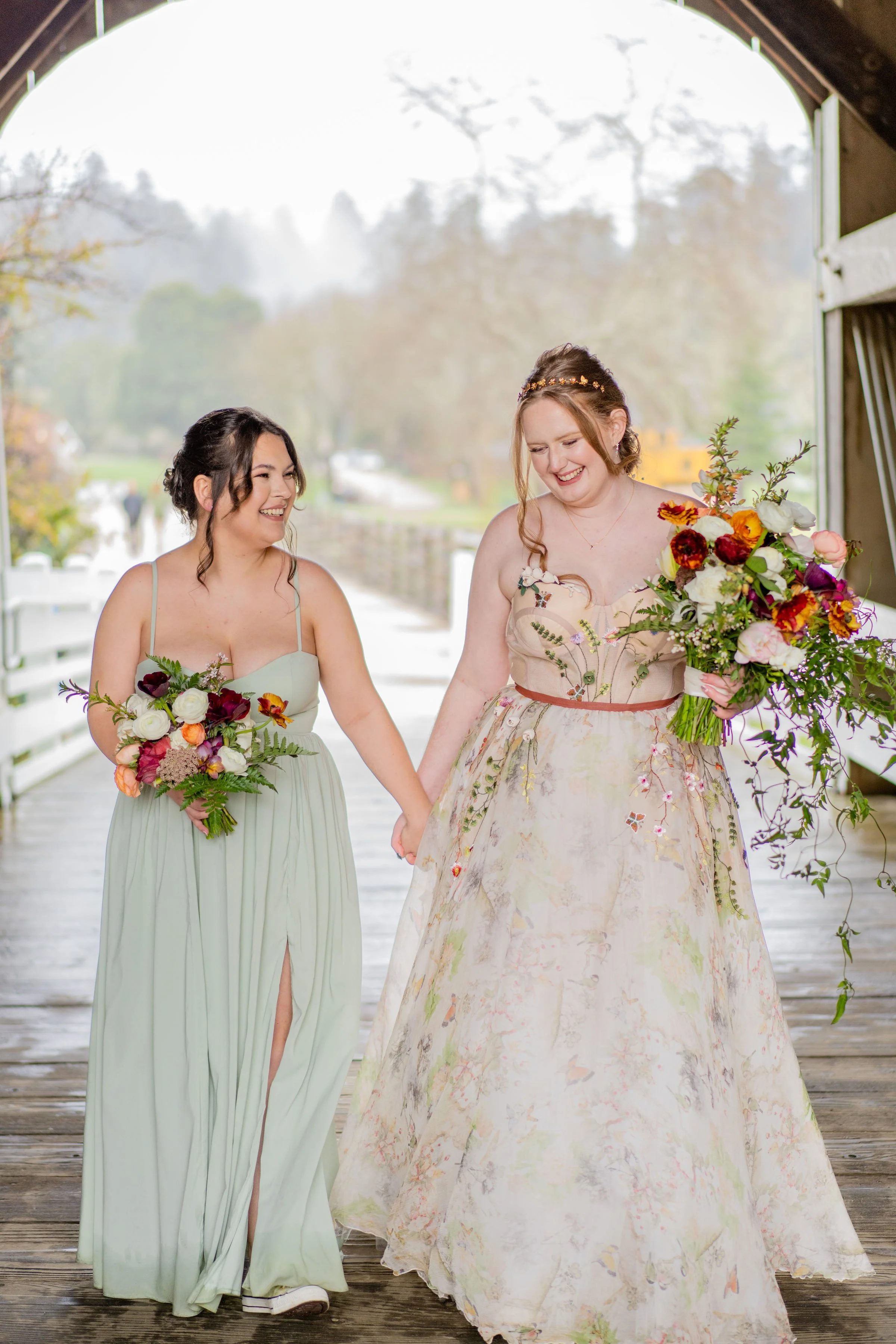 A Bride and her Maid of Honor holding lush colorful cascading bridal bouquet and bridesmaid bouquet at Roaring Camp in Santa Cruz, California. Bouquet featuring trailing jasmine vine, fall toned ranunculus, and locally grown fox glove.