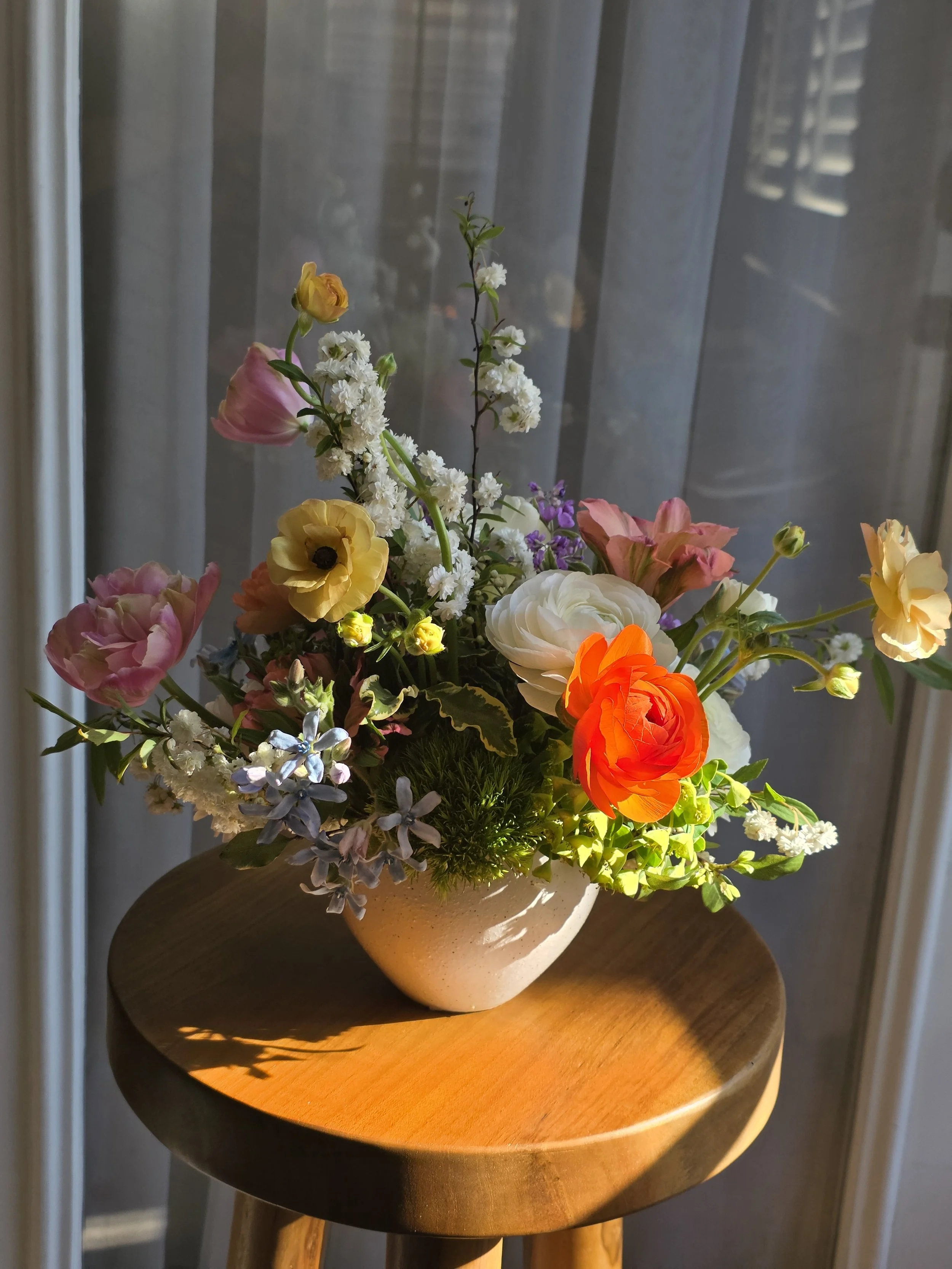 Bright mixed floral arrangement in a white ceramic vase on a wooden table with soft sunlight and sheer curtains, Petal + Salt