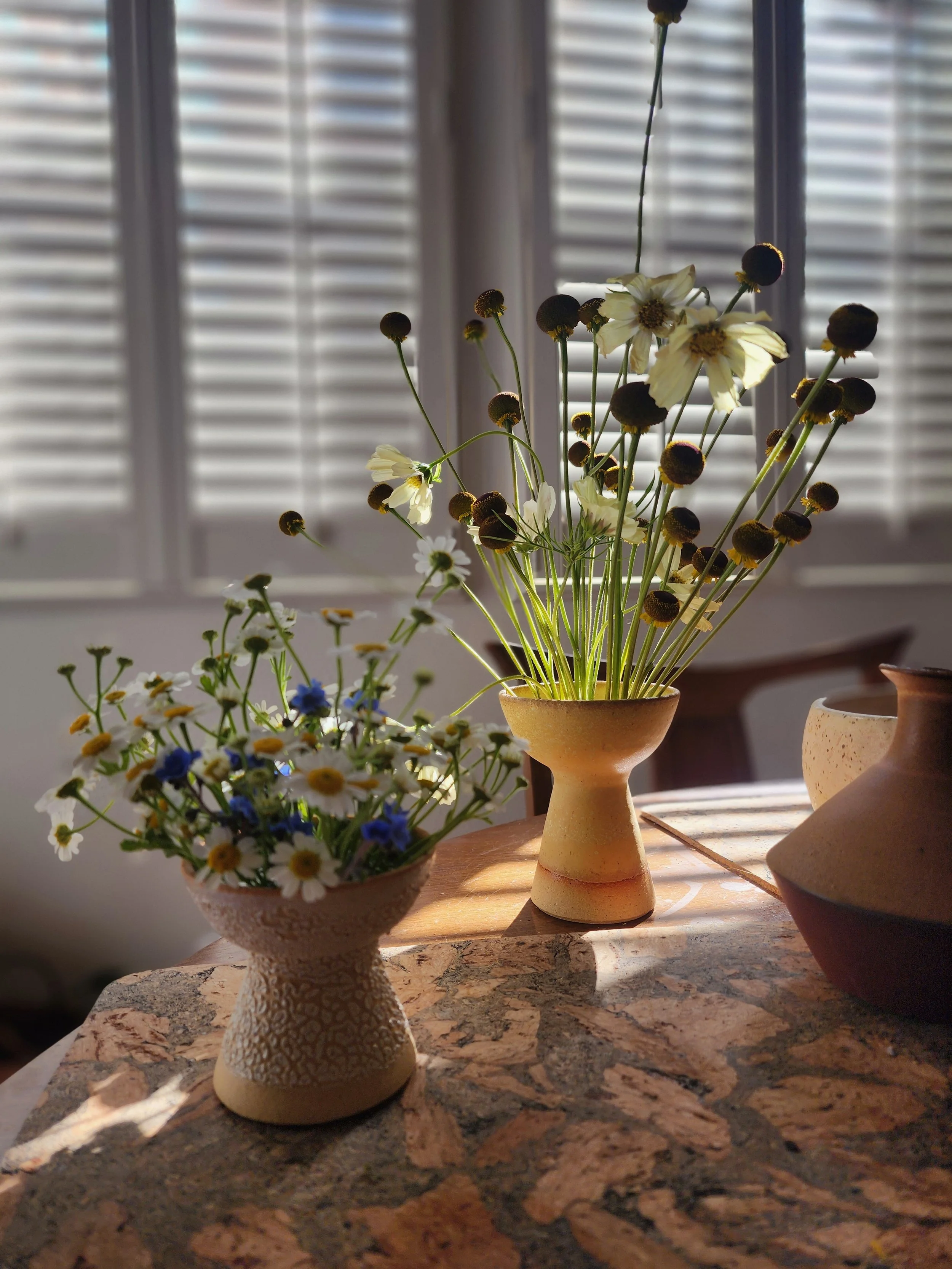 Two hand made bud vases with delicate florals on a wooden table, dramatically backlit through window blinds, Petal and Salt Floral standing order