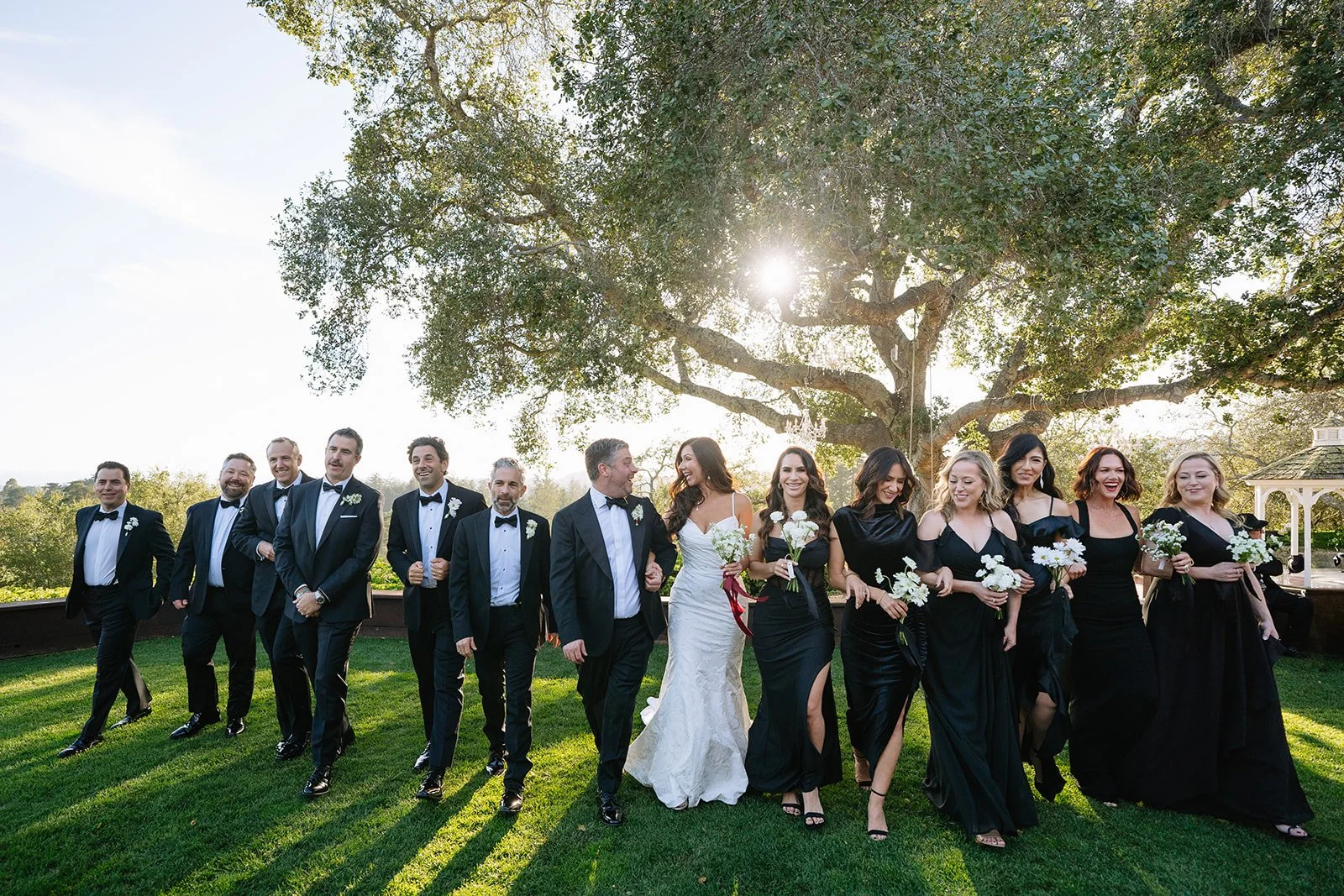Wedding party strolling under a sweeping oak at golden hour, all in black with the bride in white, florals by Petal + Salt Floral, Hollins House Santa Cruz