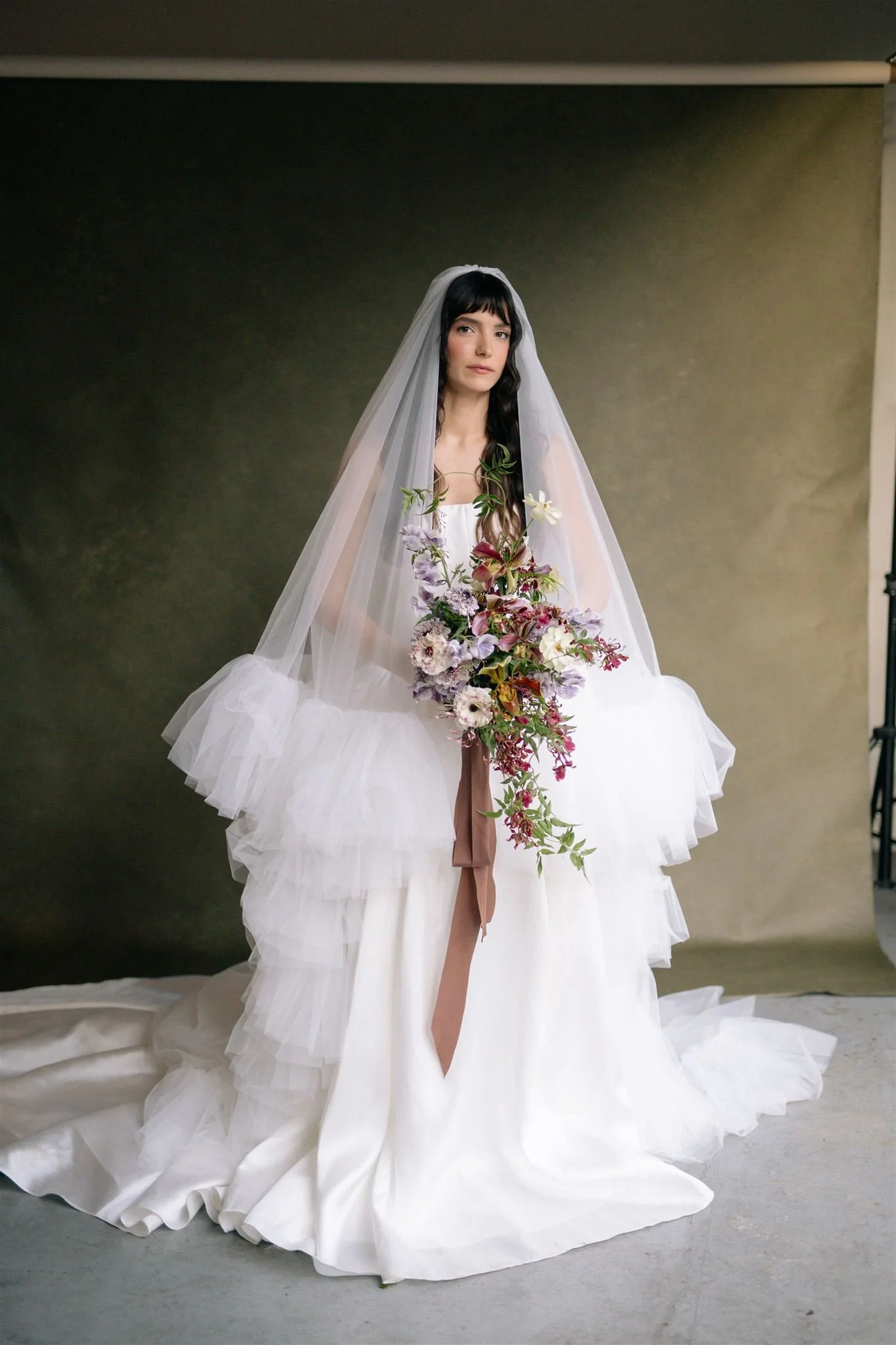 Bride with a sheer veil and couture dress holding a cascading purple, white, and green bouquet against a deep green background, Petal + Salt Floral