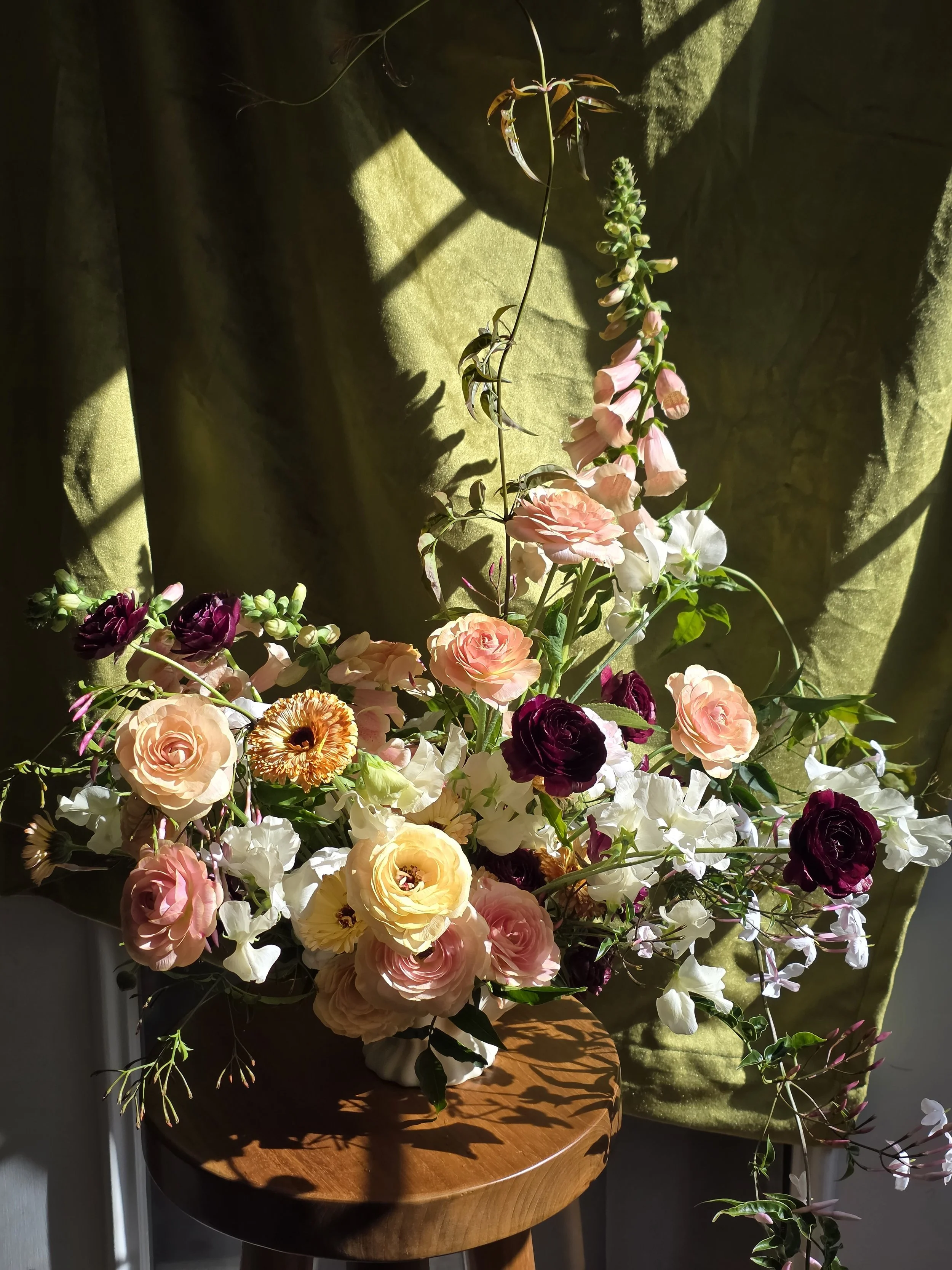 Vibrant pink, purple, yellow, and white floral arrangement on a wooden table with sunlight and a green velvet backdrop, Petal + Salt Floral