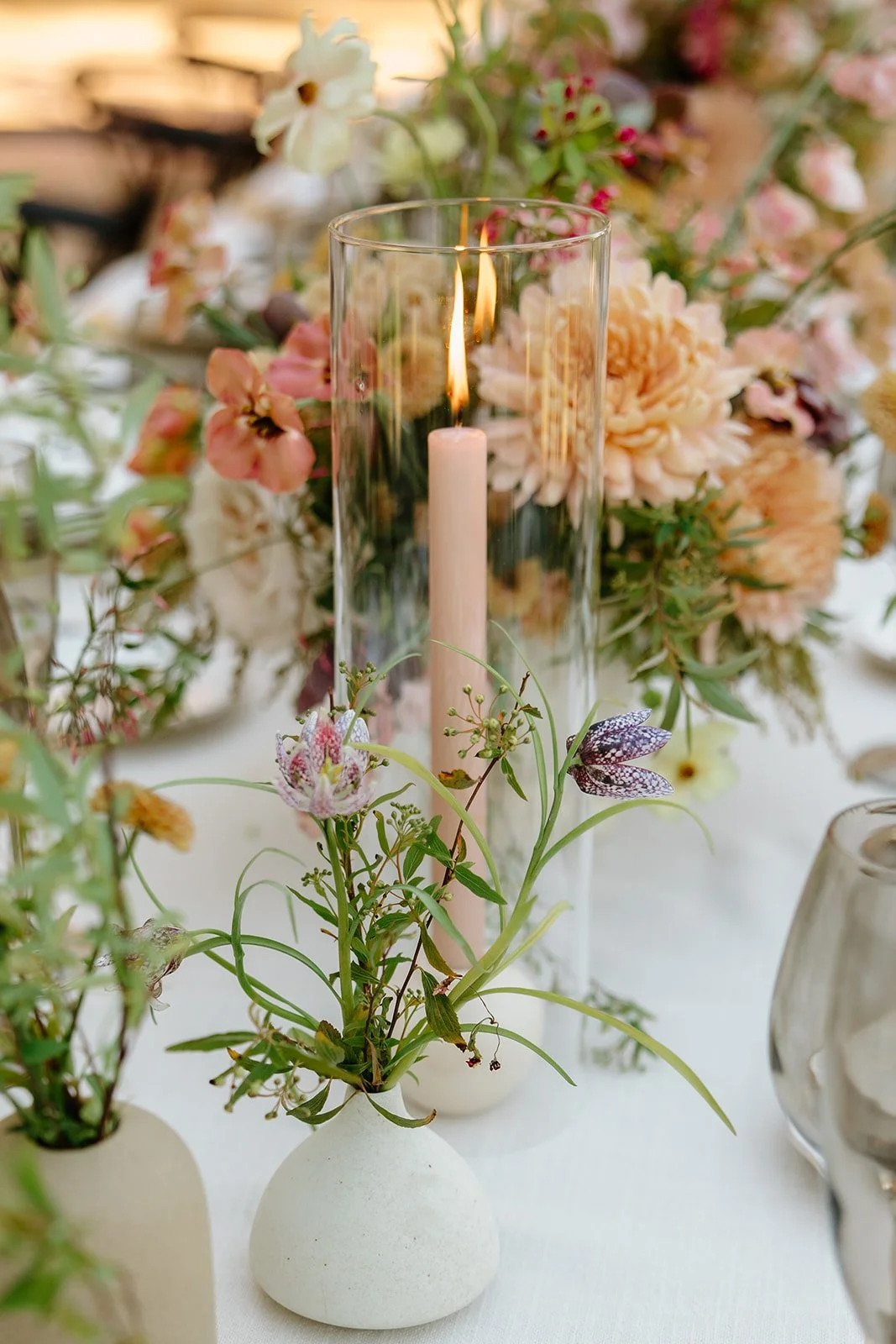 Intimate wedding tablescape with a glass pillar candle surrounded by pink, cream, and green florals in beige bud vases, Petal + Salt