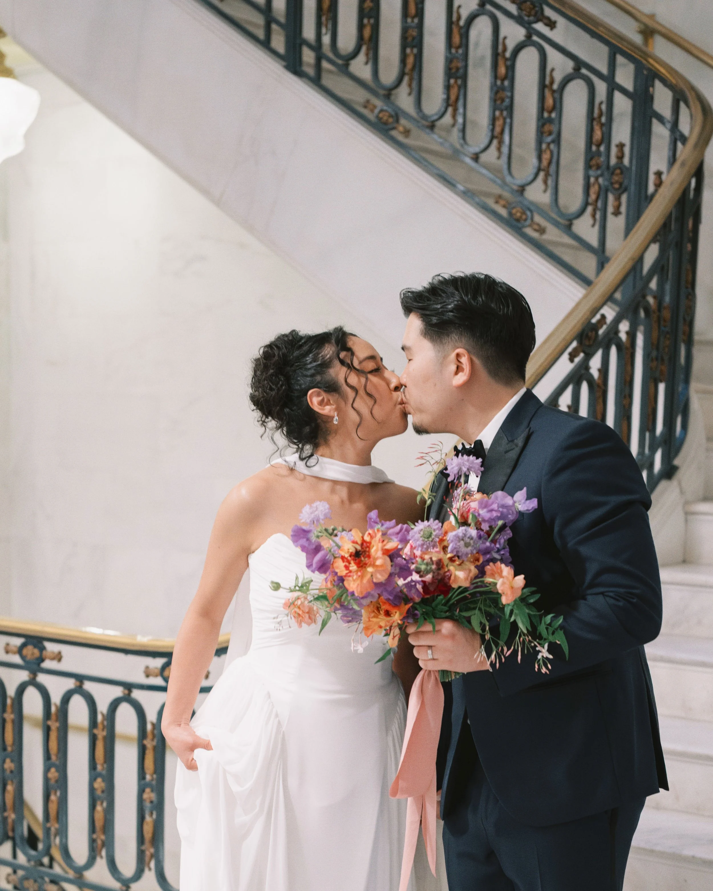 Couple sharing a kiss on a marble staircase with ornate railings, bride holding a colorful bouquet by Petal and Salt Floral