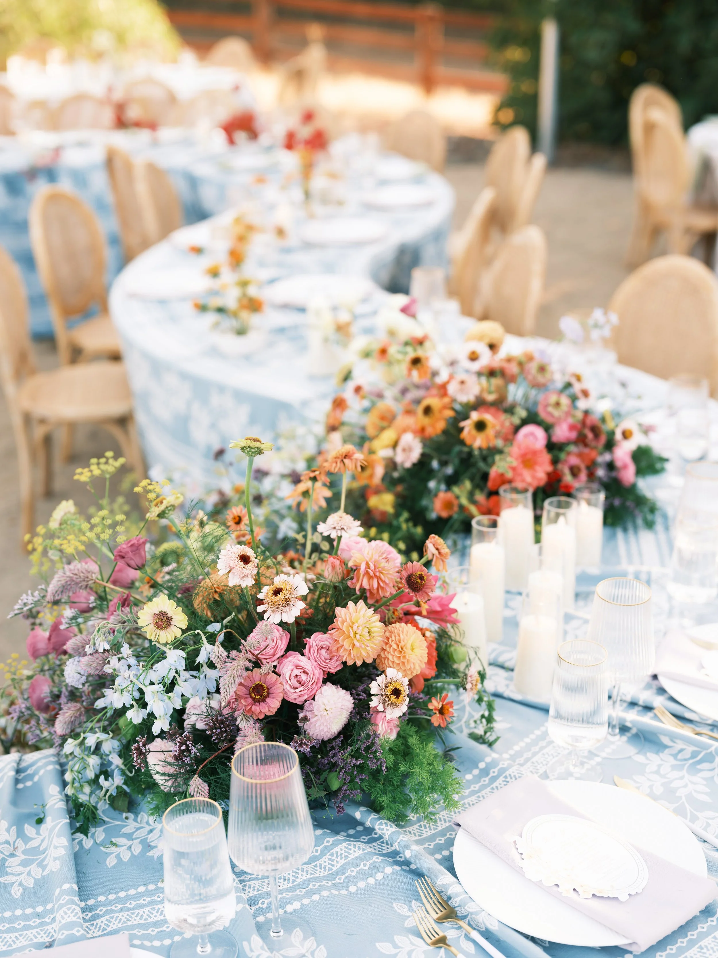 Outdoor wedding reception with a large colorful low centerpiece and tapered candles, multiple tables dressed beyond, Petal and Salt Floral
