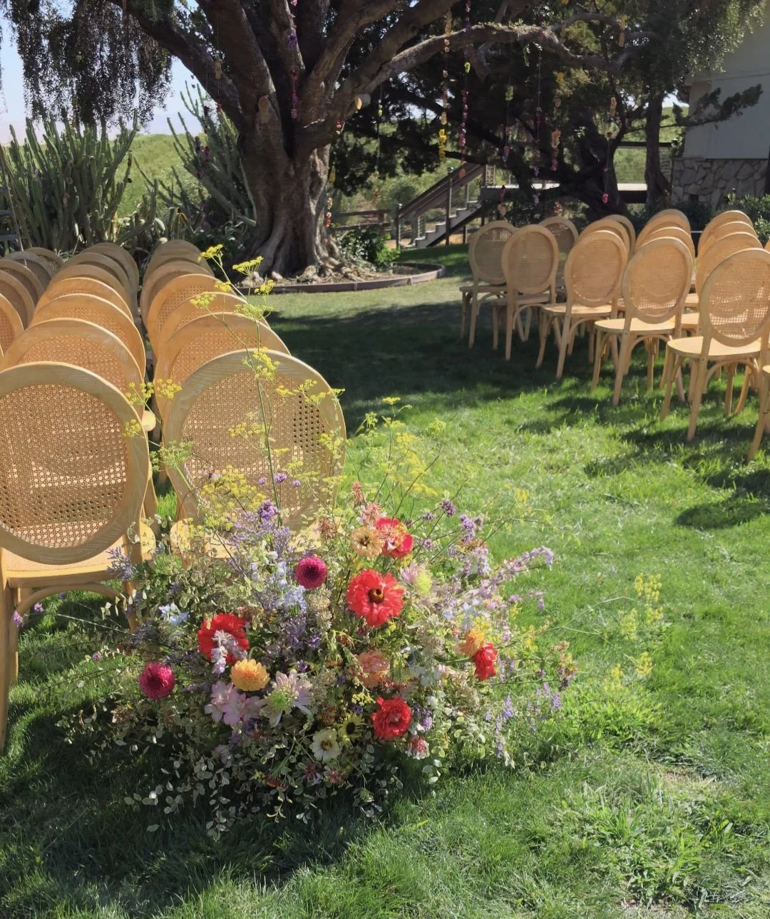Outdoor garden wedding ceremony at Olive Hill Estate with rattan bistro chairs, a flower-adorned tree, and a colorful floral arrangement in the foreground, Petal and Salt