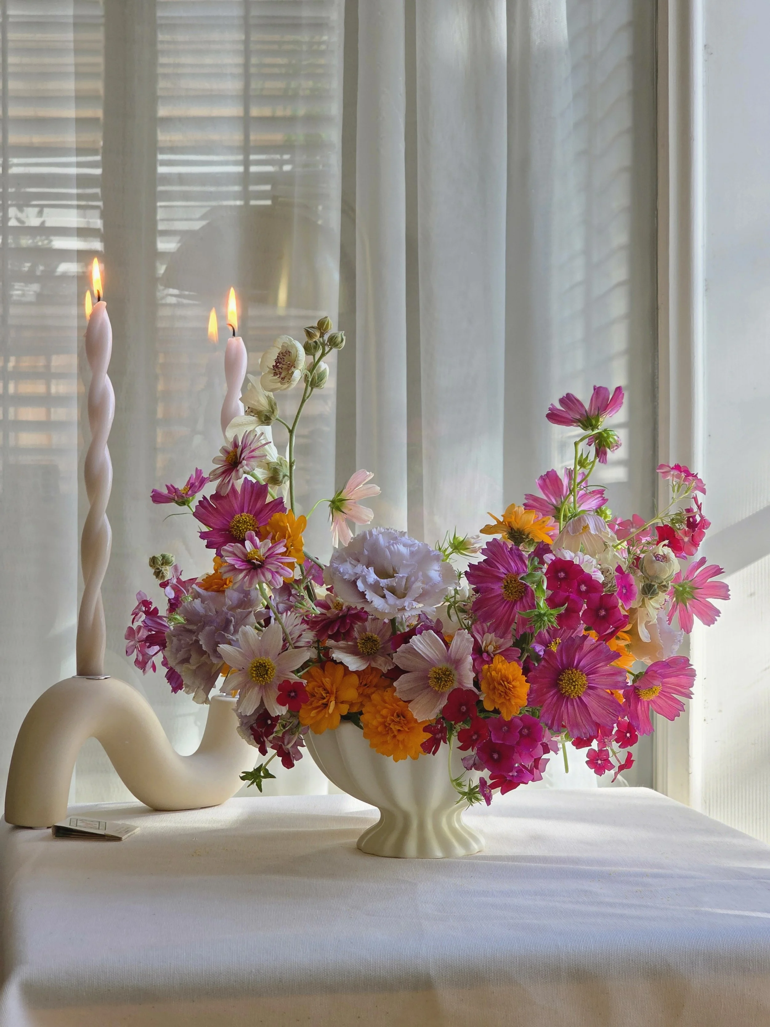 Colorful floral arrangement in a sculptural white vase, featuring cosmos, zinnias, and seasonal flowers in pink, orange, and lavender, styled with modern twisted taper candles on a linen-draped table. Wedding centerpiece pops of color, Carmel by the 