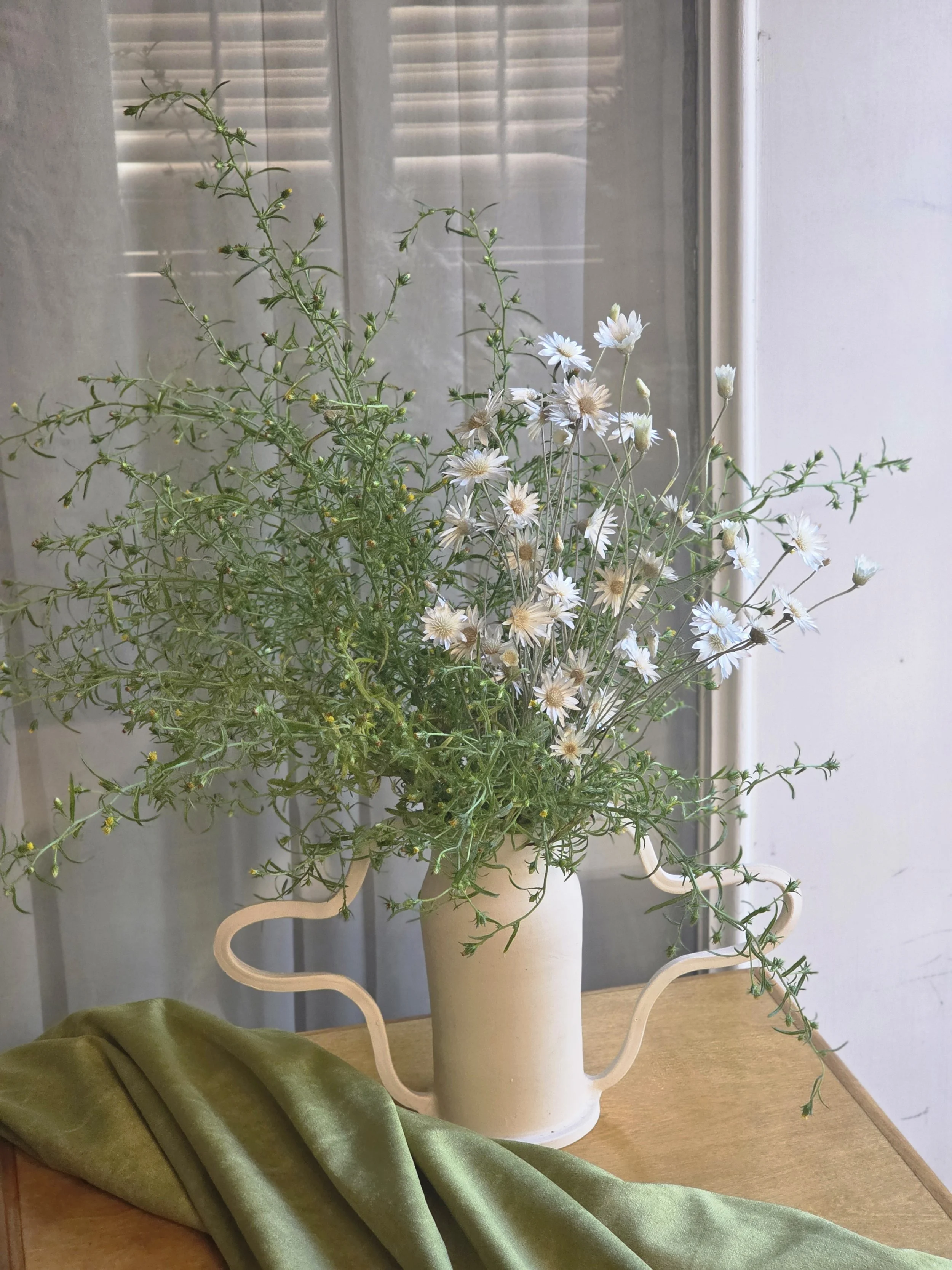 Tall white xeranthemums and native California green foliage arrangement in a hand made ceramic vase on a draped wooden table by a slatted window, Petal + Salt Floral