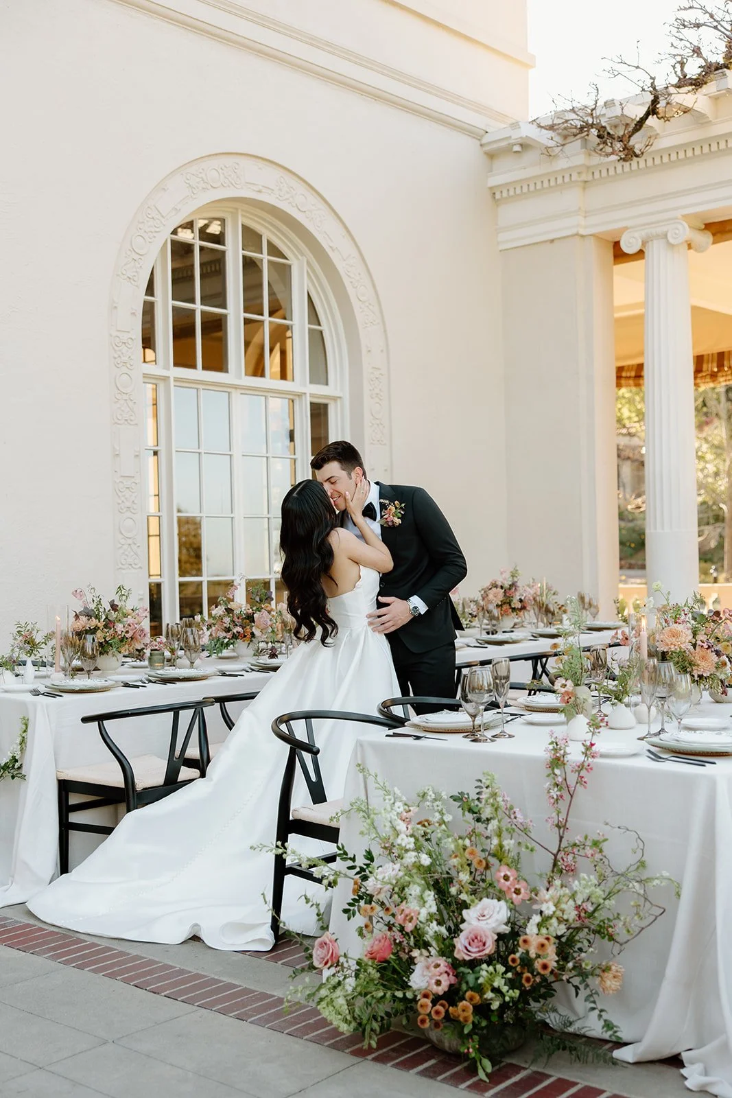 Bride and groom kiss among reception tables at Villa Montalvo in Saratoga, California, surrounded by romantic garden-style arrangements of roses, dahlias, and seasonal blooms in soft pinks and creams. Large ground arrangement bookending reception tab