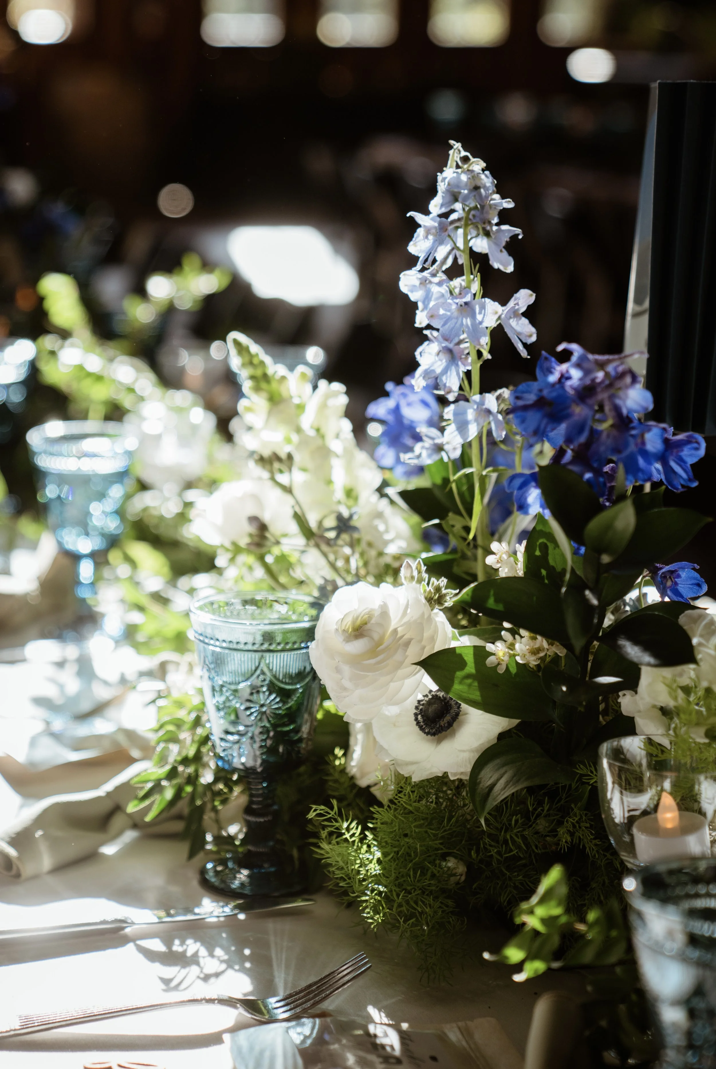 Elegant floral centerpiece with white roses, blue delphiniums, and greenery on a tablescape with candles at Roaring Camp in Santa Cruz, California. 