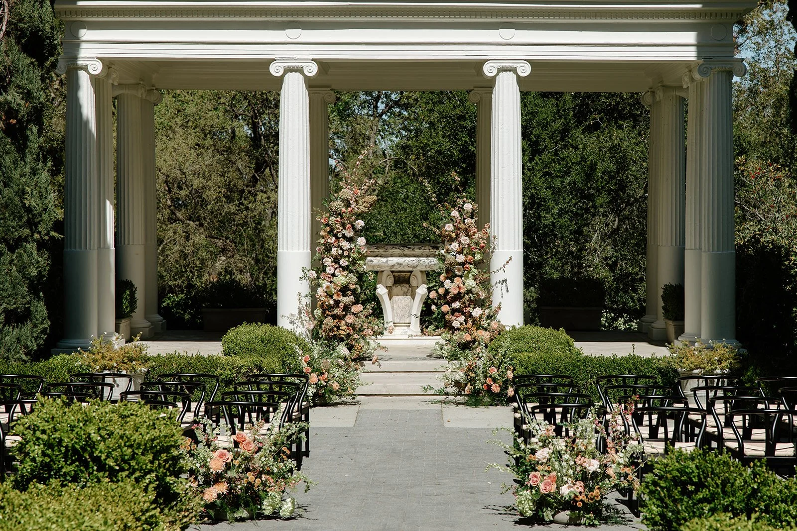 Wedding ceremony at Villa Montalvo in Saratoga, California, featuring a classical white pavilion decorated with lush floral arch of roses, dahlias, and hydrangea in soft romantic tones, with ground arrangements lining the aisle.