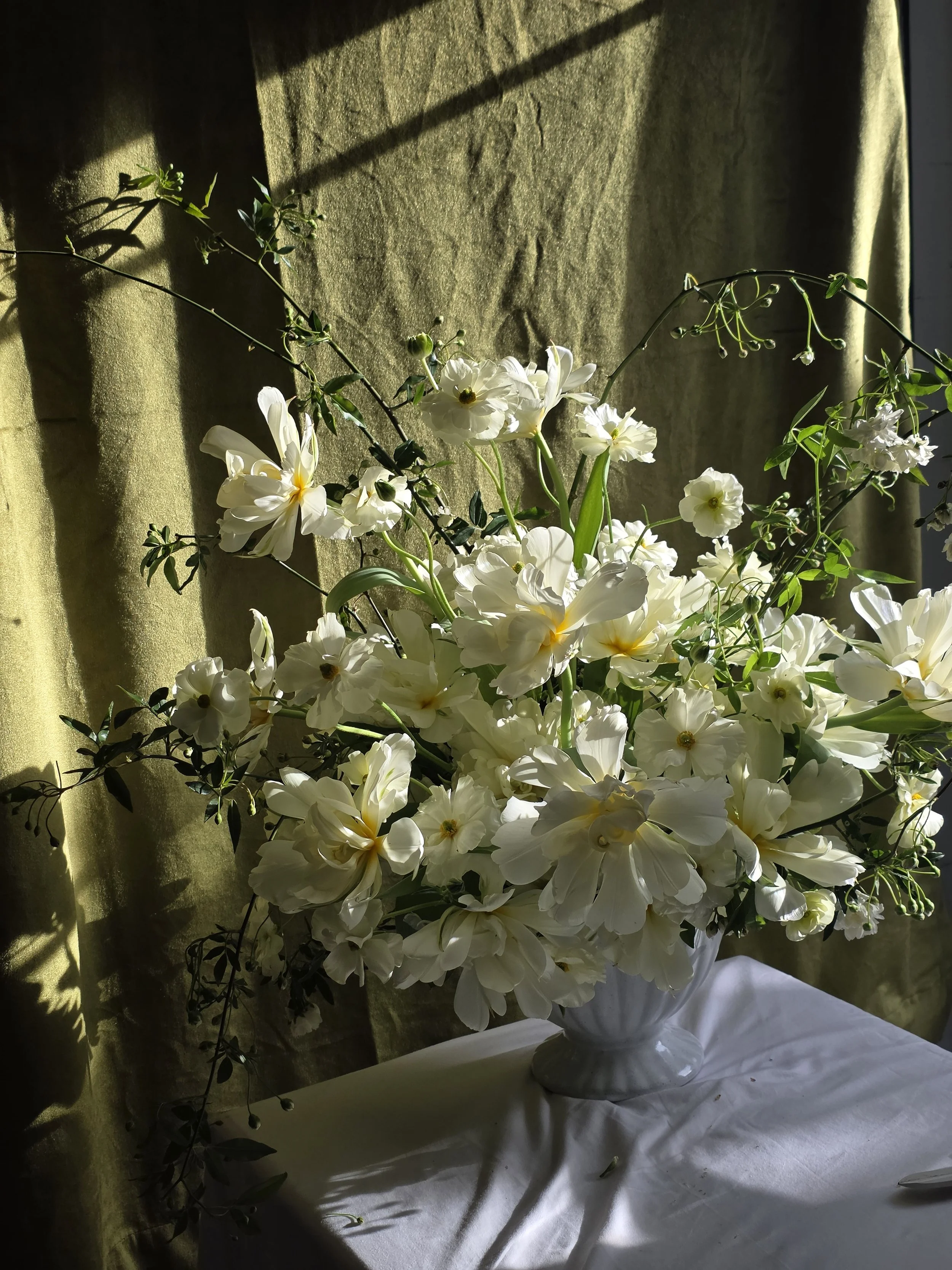 Voluminous all-butter yellow floral arrangement with exotic emperor tuplips, butterfly ranunculus, and lady banks roses in a white vase at a sunlit reception table, Petal + Salt Floral