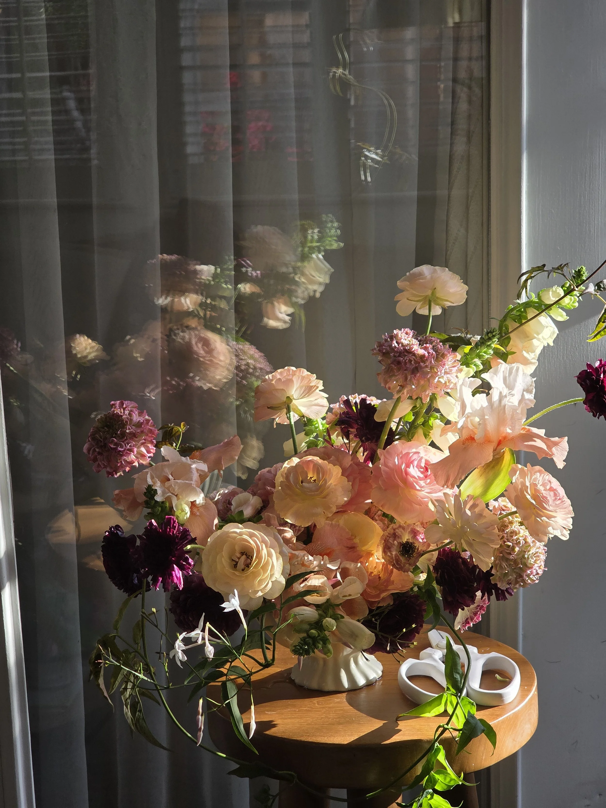 Peach, pink, white, and burgundy floral arrangement made mostly with locally grown ranunculus, foxglove, and bearded irises in a Camilia Supply compote by a sheer-curtained window, standing order florals by Petal + Salt