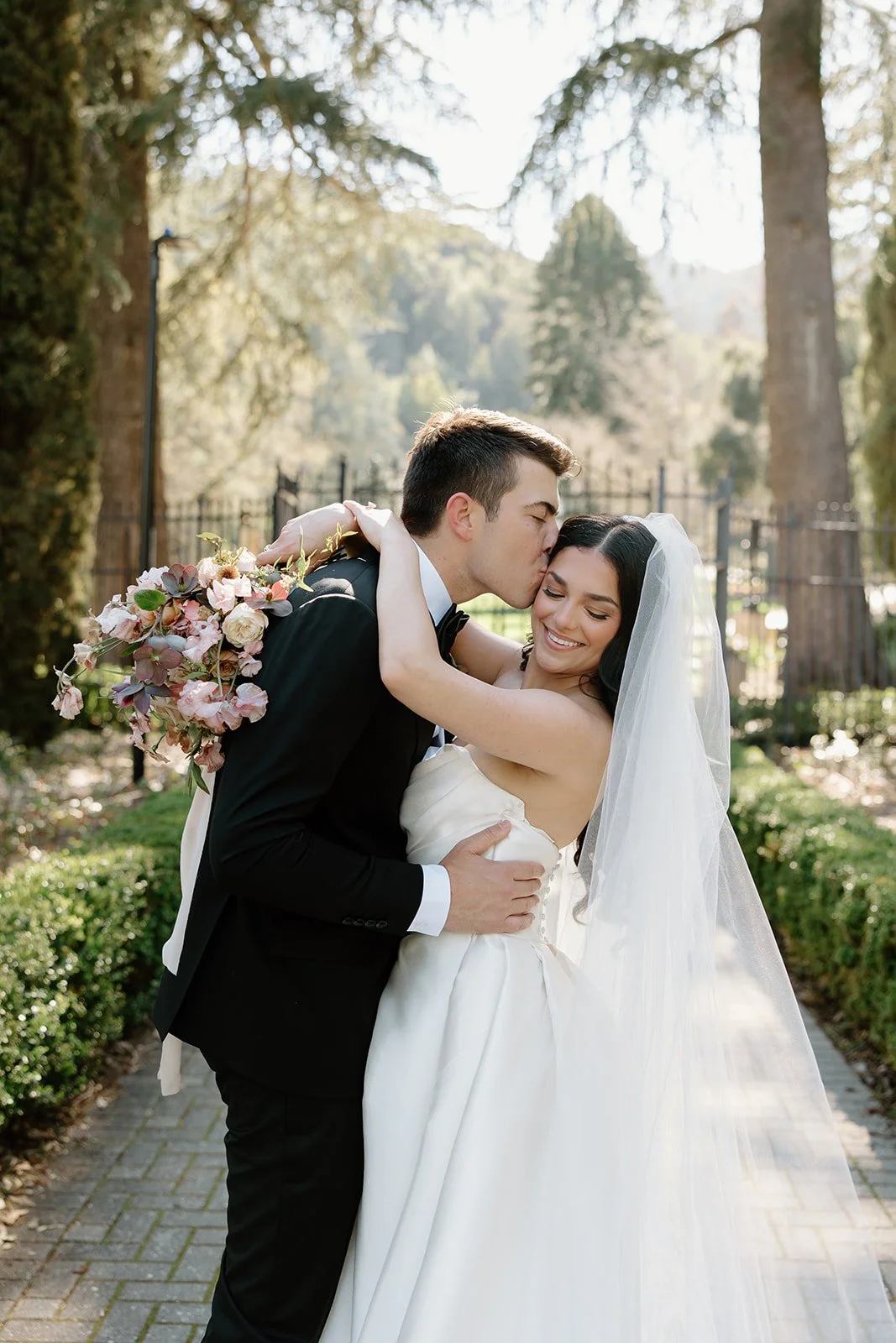 Newlywed couple at Villa Montalvo in Saratoga, California, sharing a joyful embrace. The bride holds a romantic bouquet of garden roses and sweet peas in soft pastel tones.