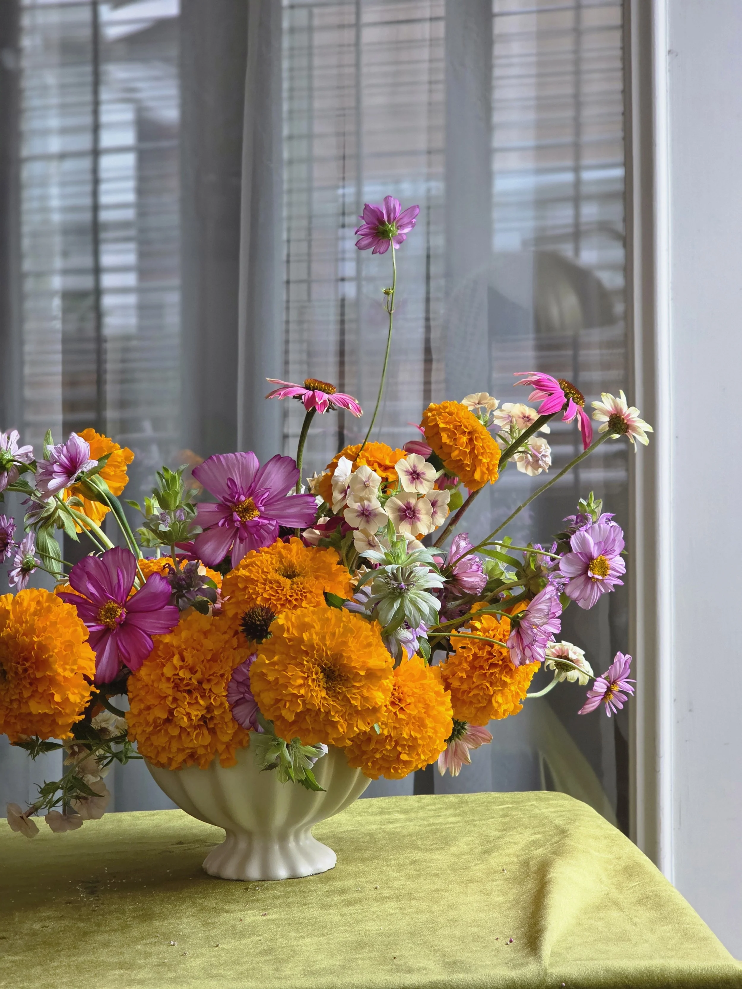 Vivid pink, orange, and white arrangement in a white ceramic Camilia Supply vase on a green cloth surface by a sheer window, Petal and Salt Floral