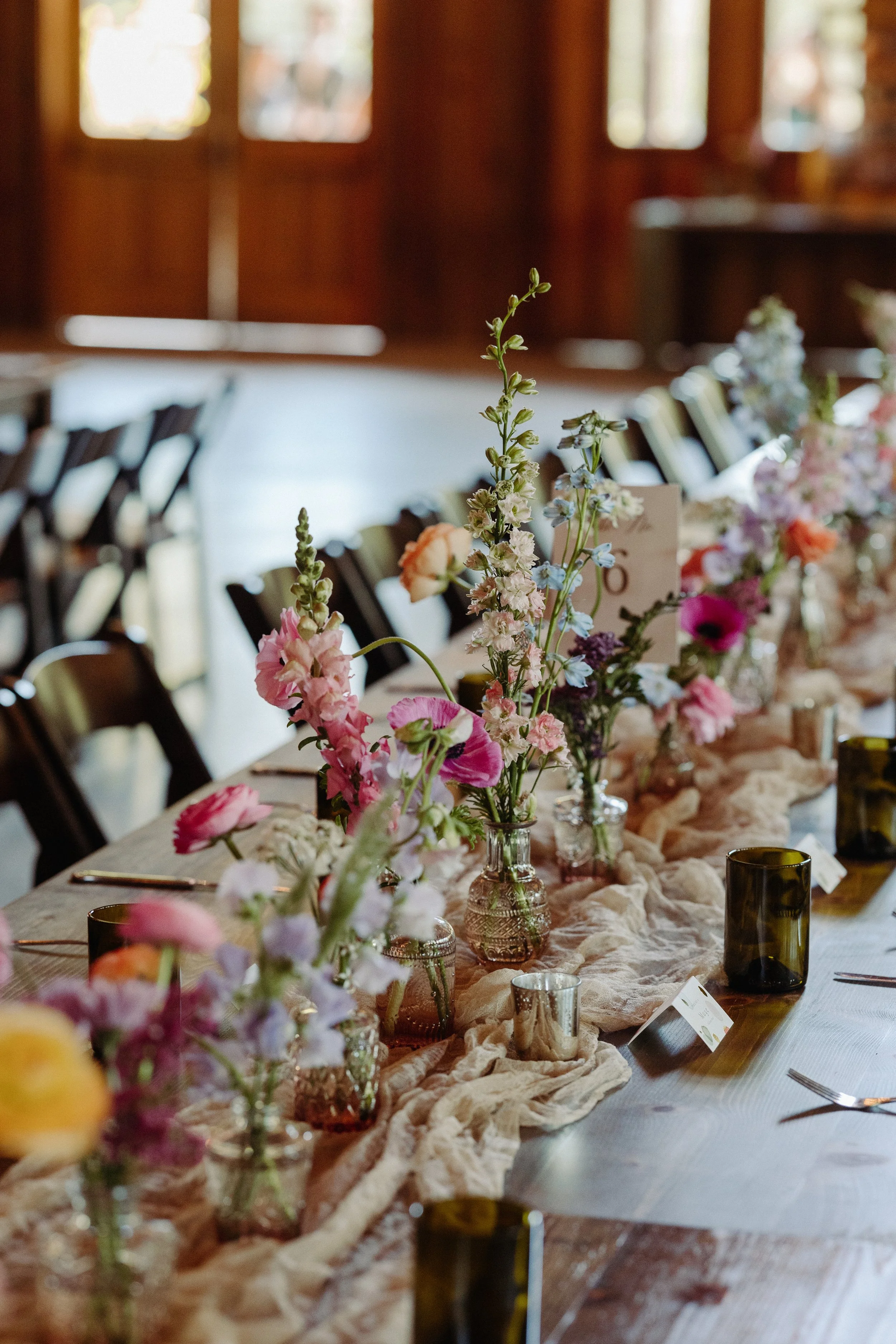 A decorated table with colorful flower arrangements in glass vases, set with tableware, and draped with a light fabric runner, Wedding Reception flowers. Santa Cruz California wedding reception at Roaring Camp. 