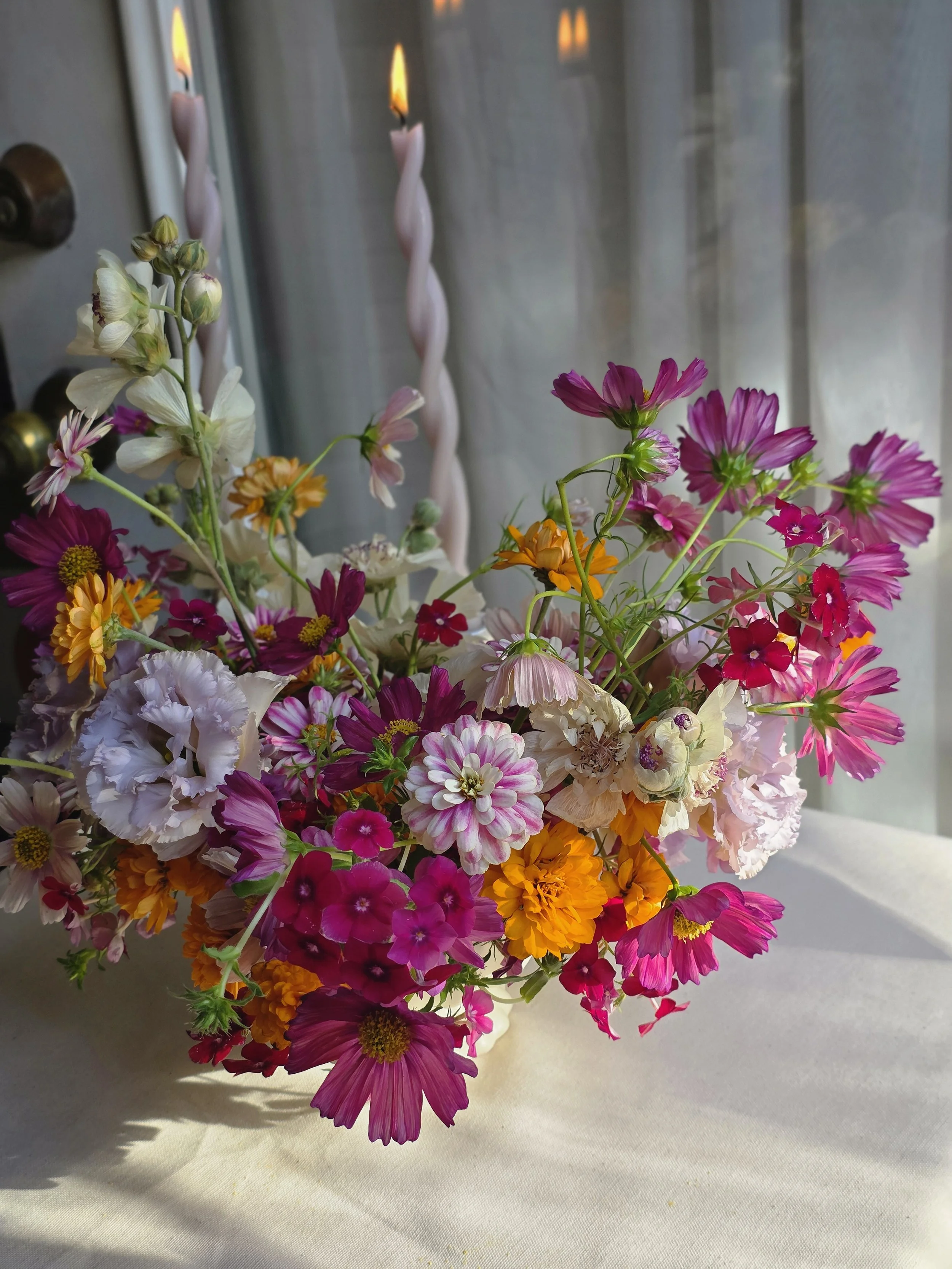 Vibrant mixed floral arrangement alongside twisted pink taper candles by a sheer-curtained window, Petal and Salt Floral