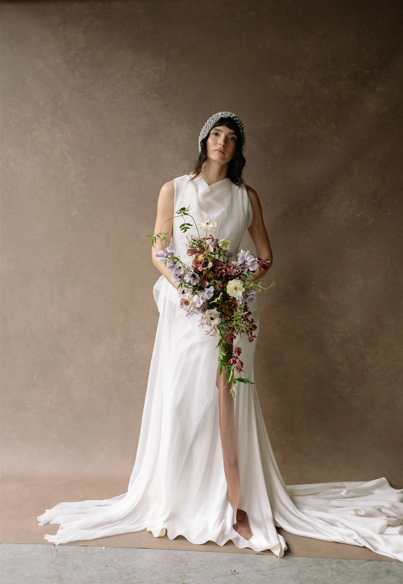 Bride in a fitted white gown holding a large lush bouquet against a warm neutral Chasing Stone backdrop, Petal and Salt Floral