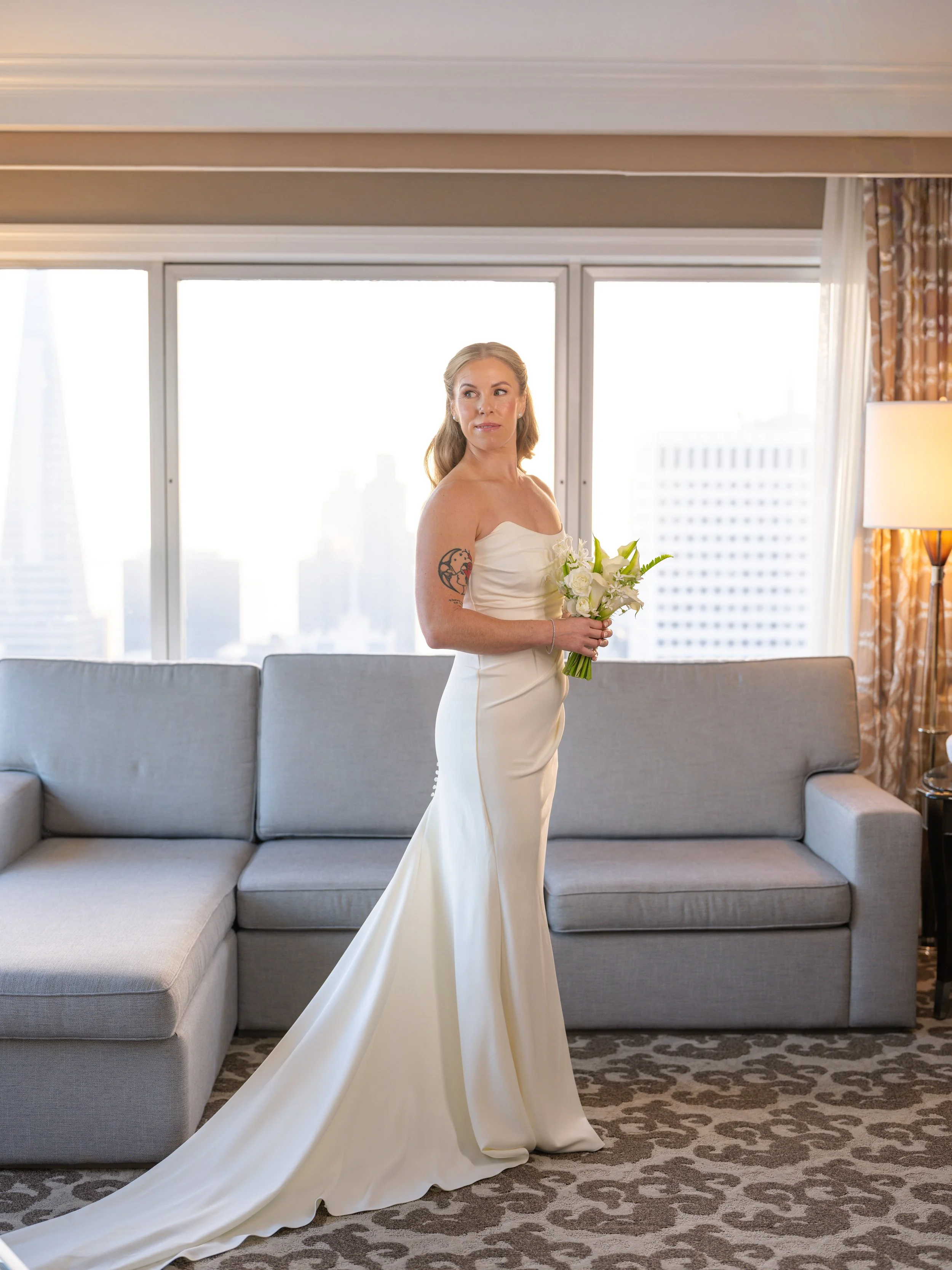  Bride in a strapless gown holding her delicate all white birdal bouquet with a city skyline visible through floor-to-ceiling windows in her Fairmont San Francisco hotel suite, Petal + Salt Floral