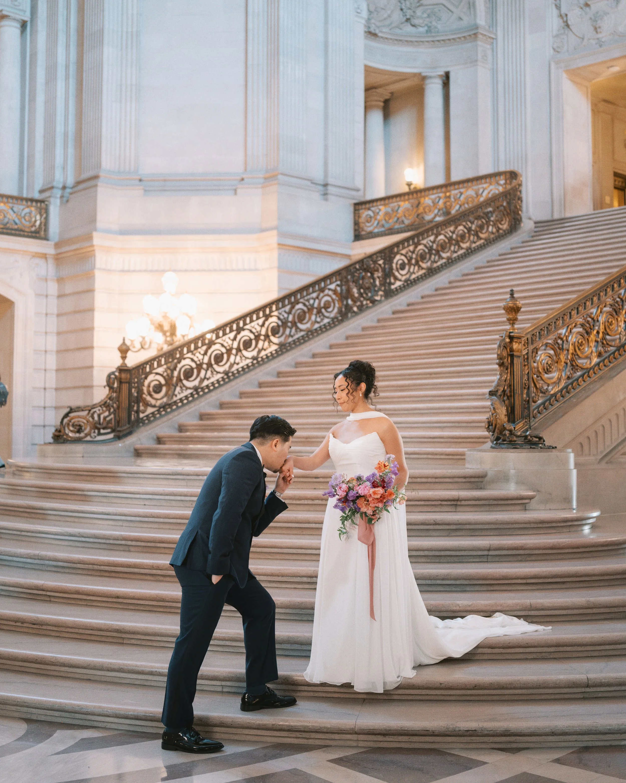 Groom kissing bride's hand on a grand marble staircase in the historic San Francisco City Hall wedding venue, bouquet by Petal + Salt Floral, Bay Area