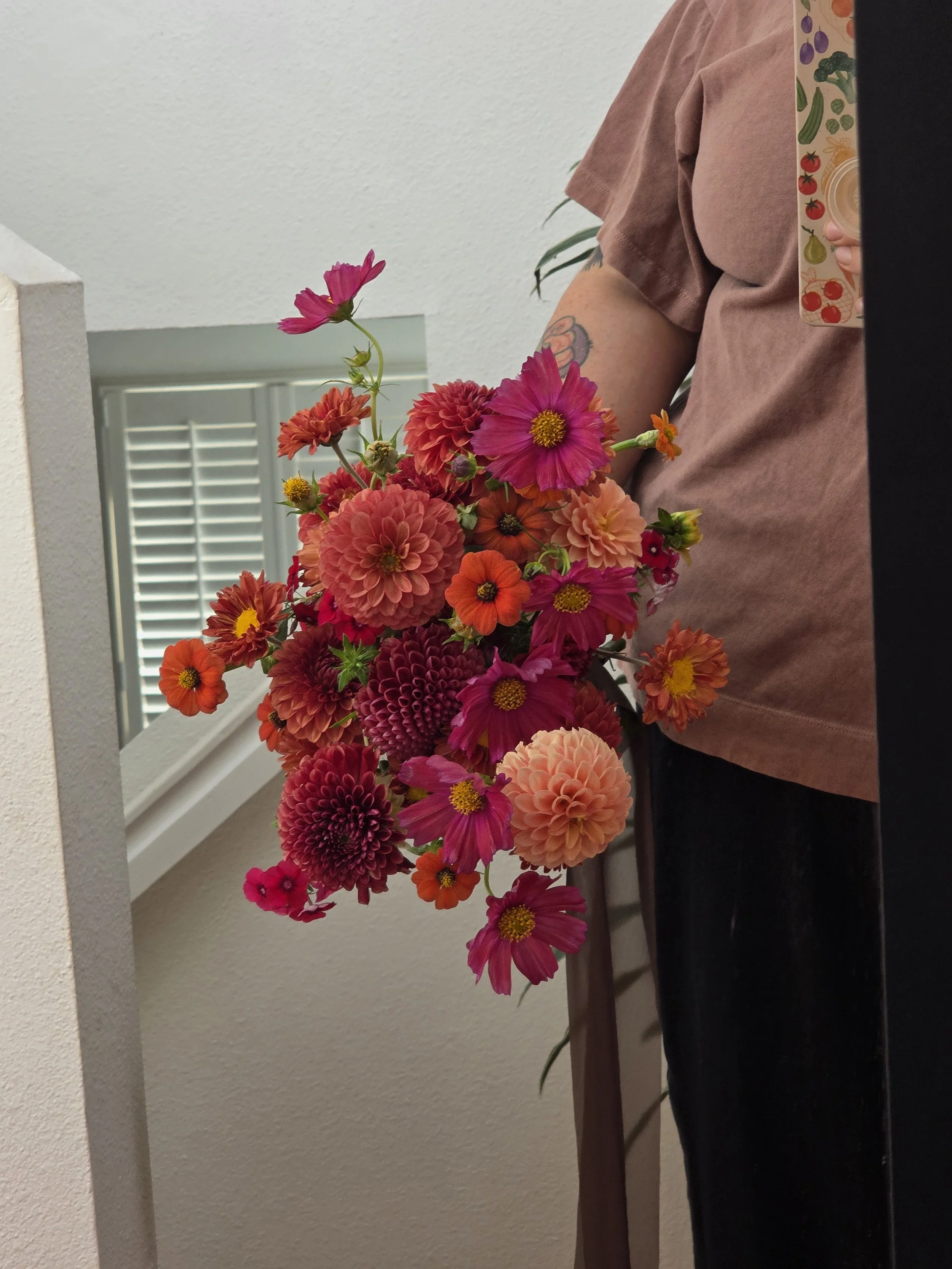 Hand-tied bouquet of pink, orange, red, and peach blooms held near a mirror, designed by Petal and Salt Floral