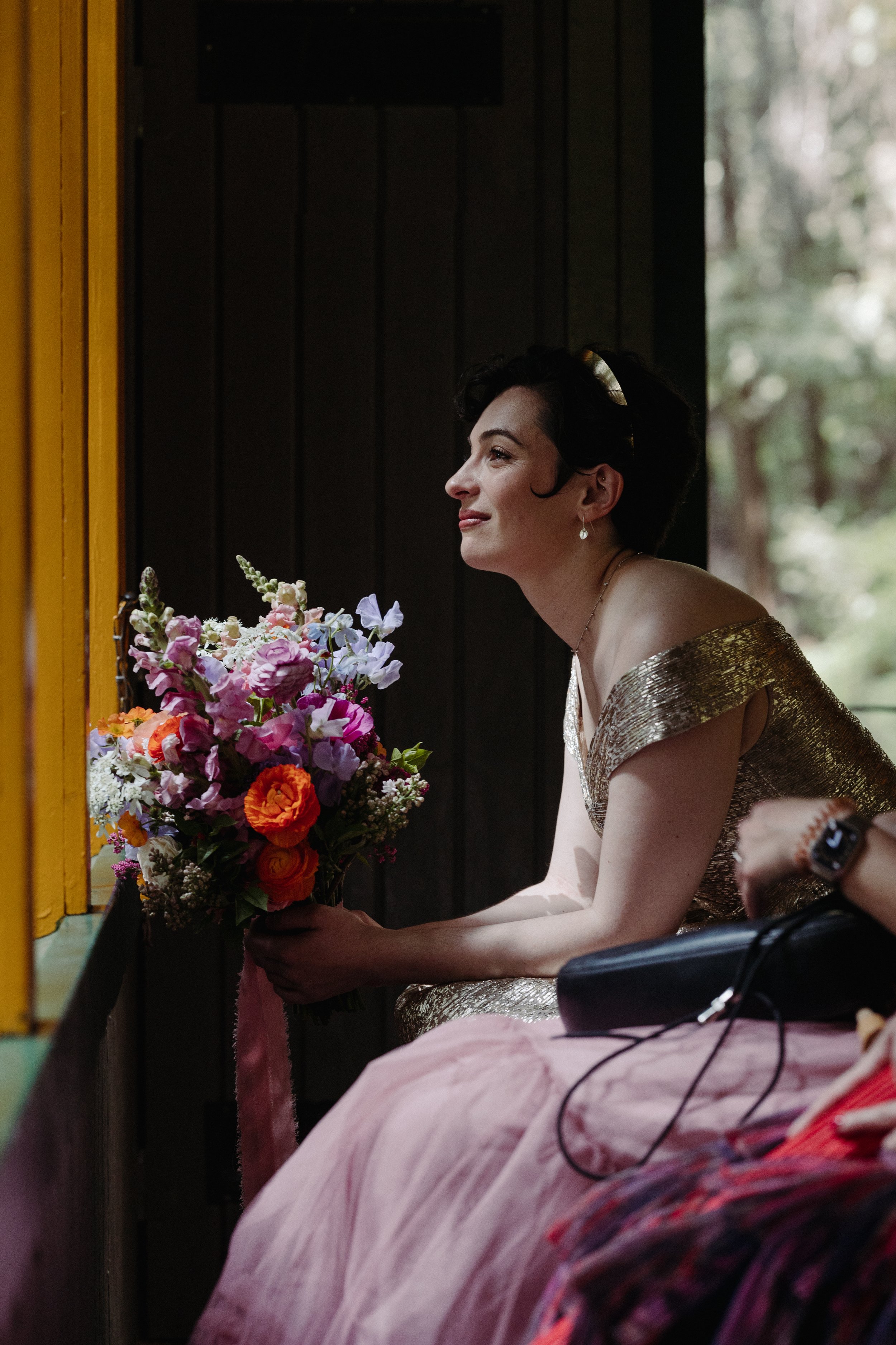 Bride riding the Roaring Camp Train up to her Wedding Ceremony holding her colorful vibrant Bridal Bouquet featuring ranunculus, delphinium, freesia, and sweet pea flowers. Santa Cruz Mountain Wedding. Wedding among the Redwoods. 